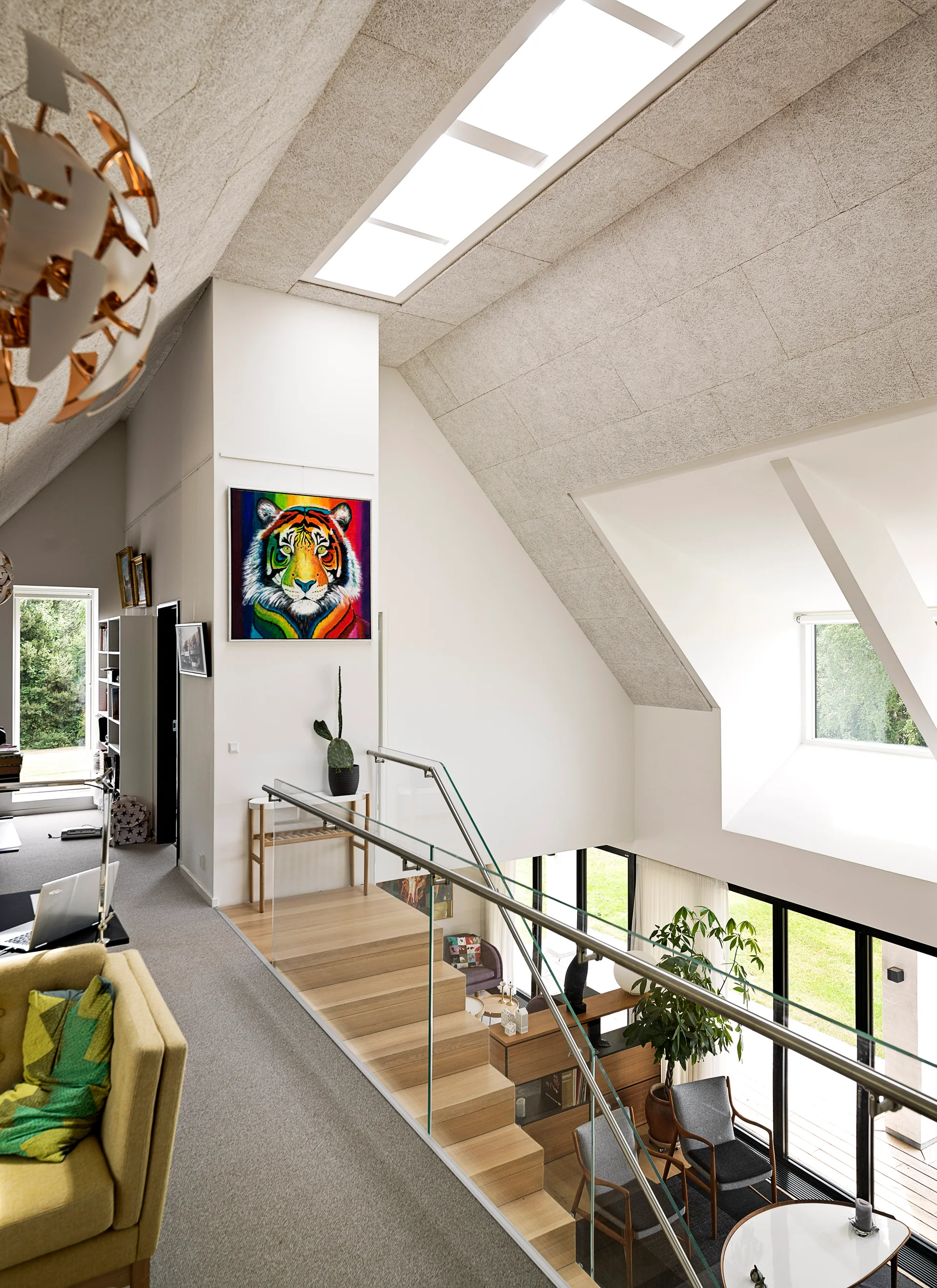 A contemporary loft space with a wooden staircase, glass railing, textured sloped ceiling with a skylight, and a vibrant tiger painting on the wall.