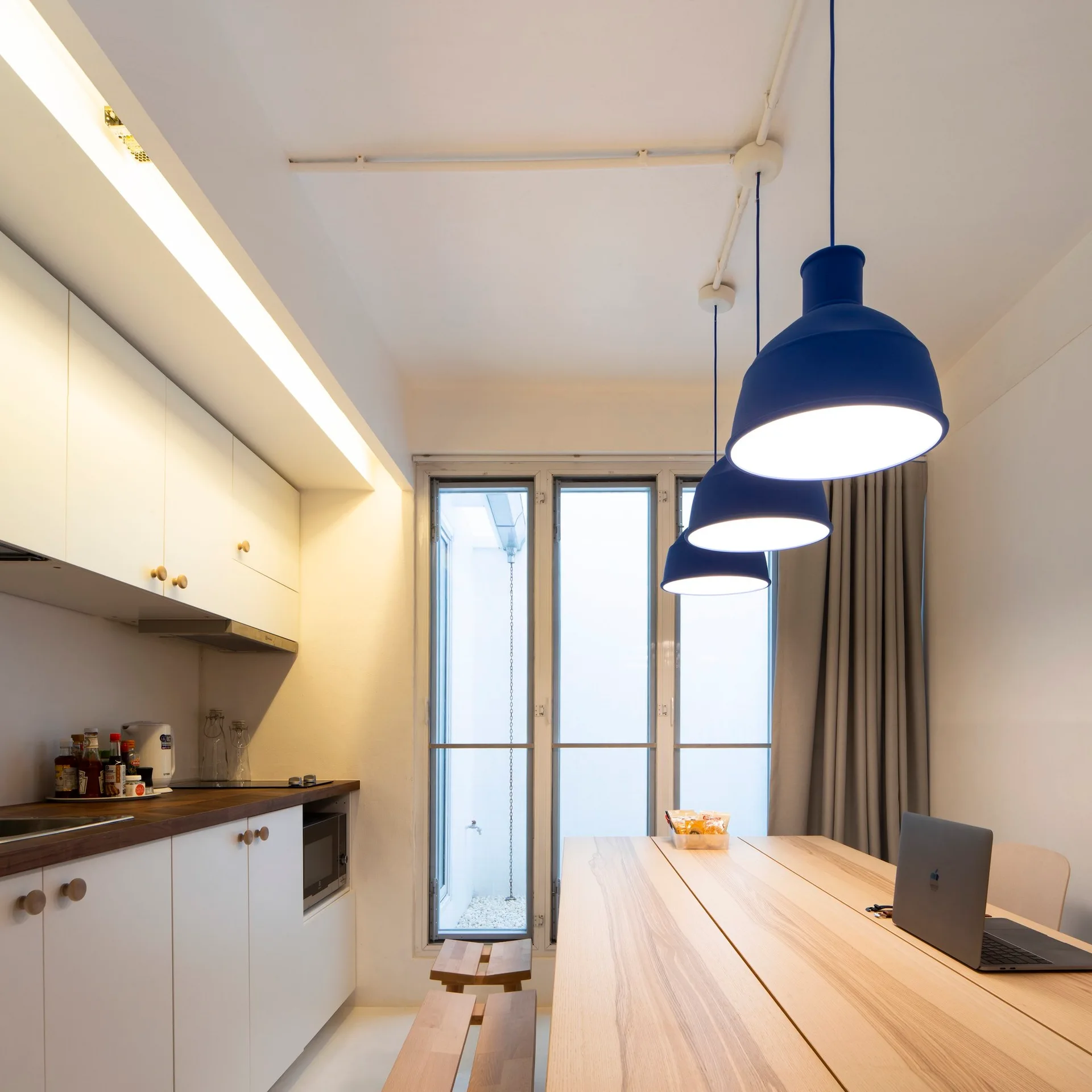 A minimalist white kitchen and dining area featuring a light wood table, simple wooden benches, and three blue pendant lights hanging overhead.