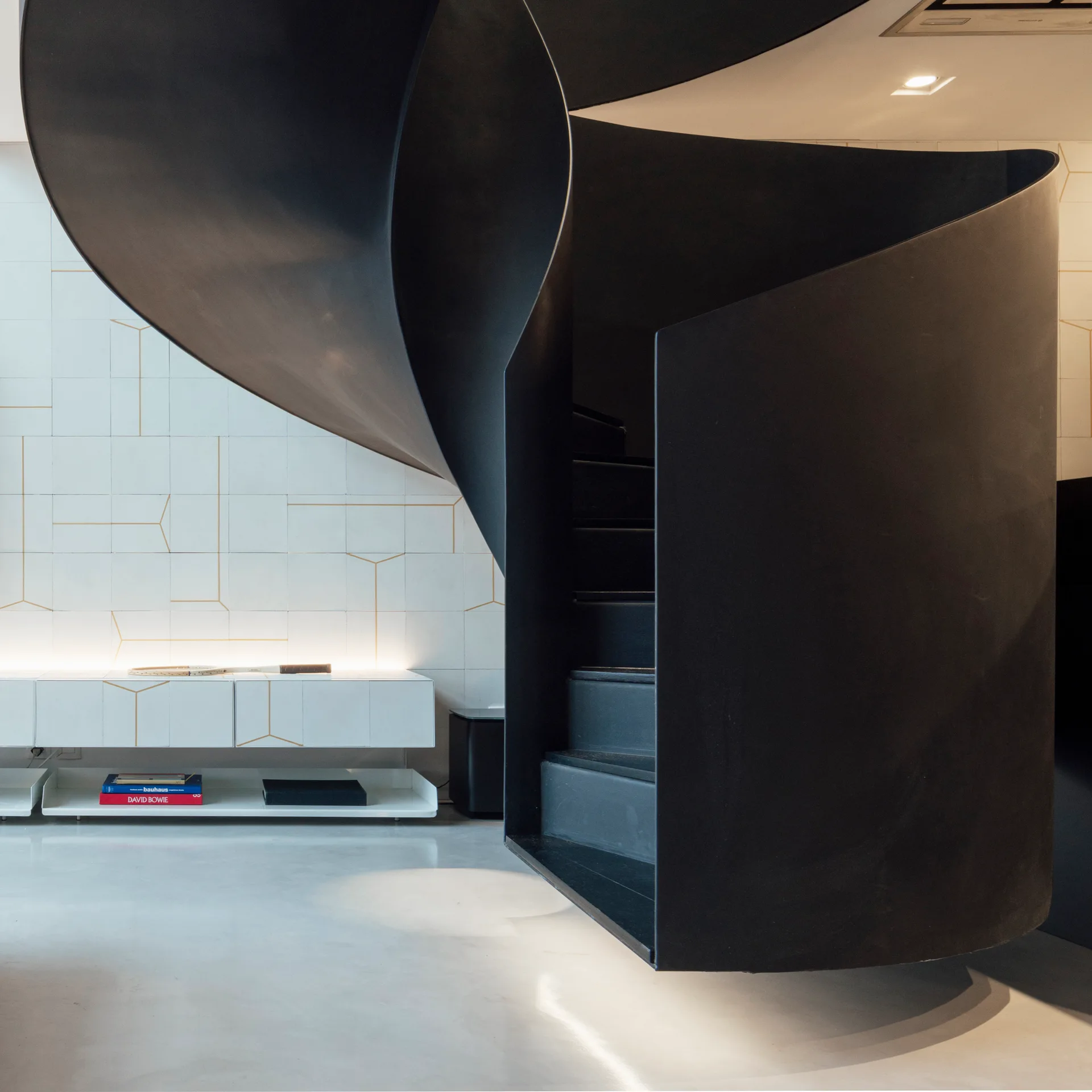 A black metal spiral staircase curves upward next to a white tiled wall detailed with gold geometric lines and a low floating console.