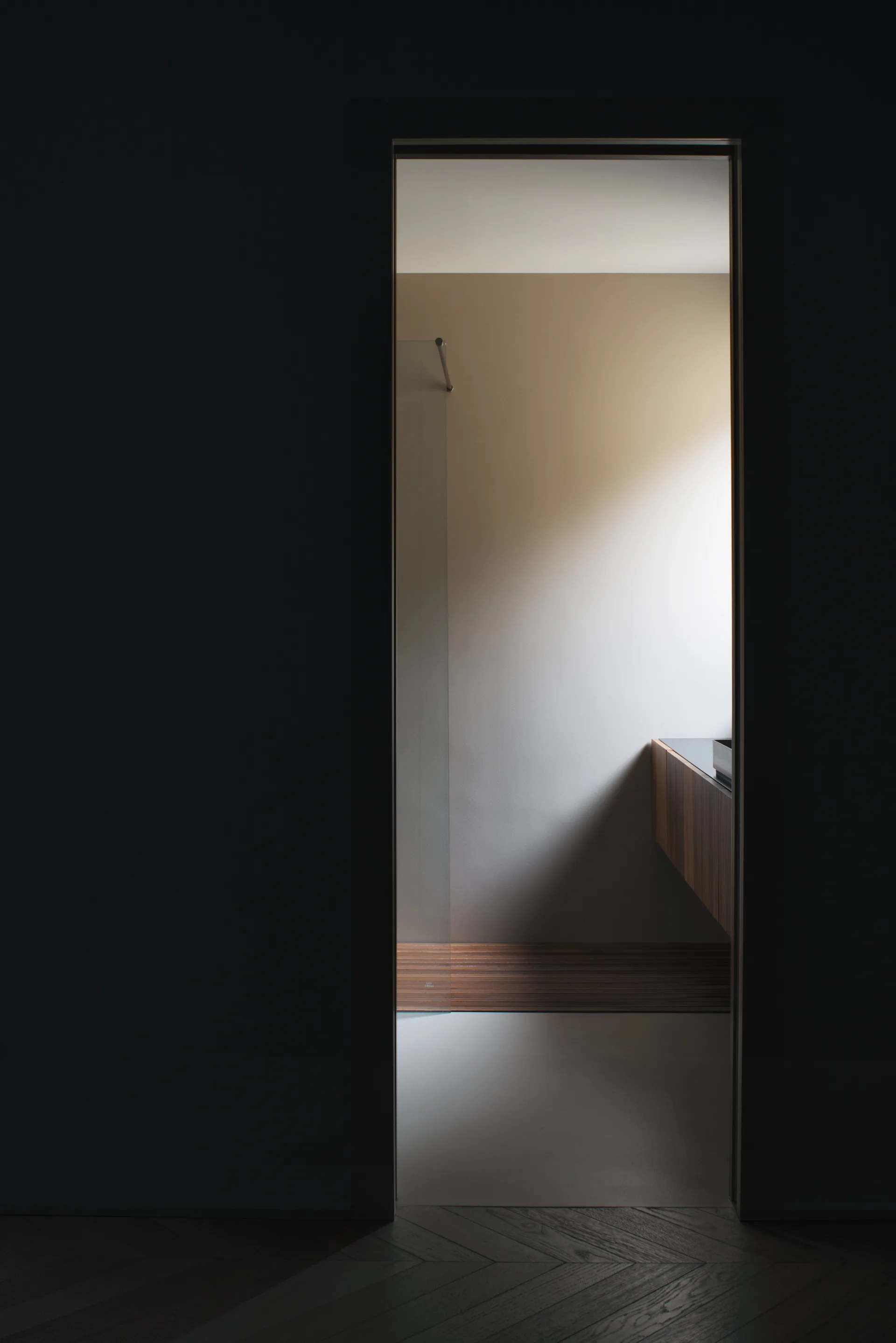 A view through a dark doorway into a minimalist bathroom featuring a floating wood slatted vanity, a glass shower partition, and matte walls.
