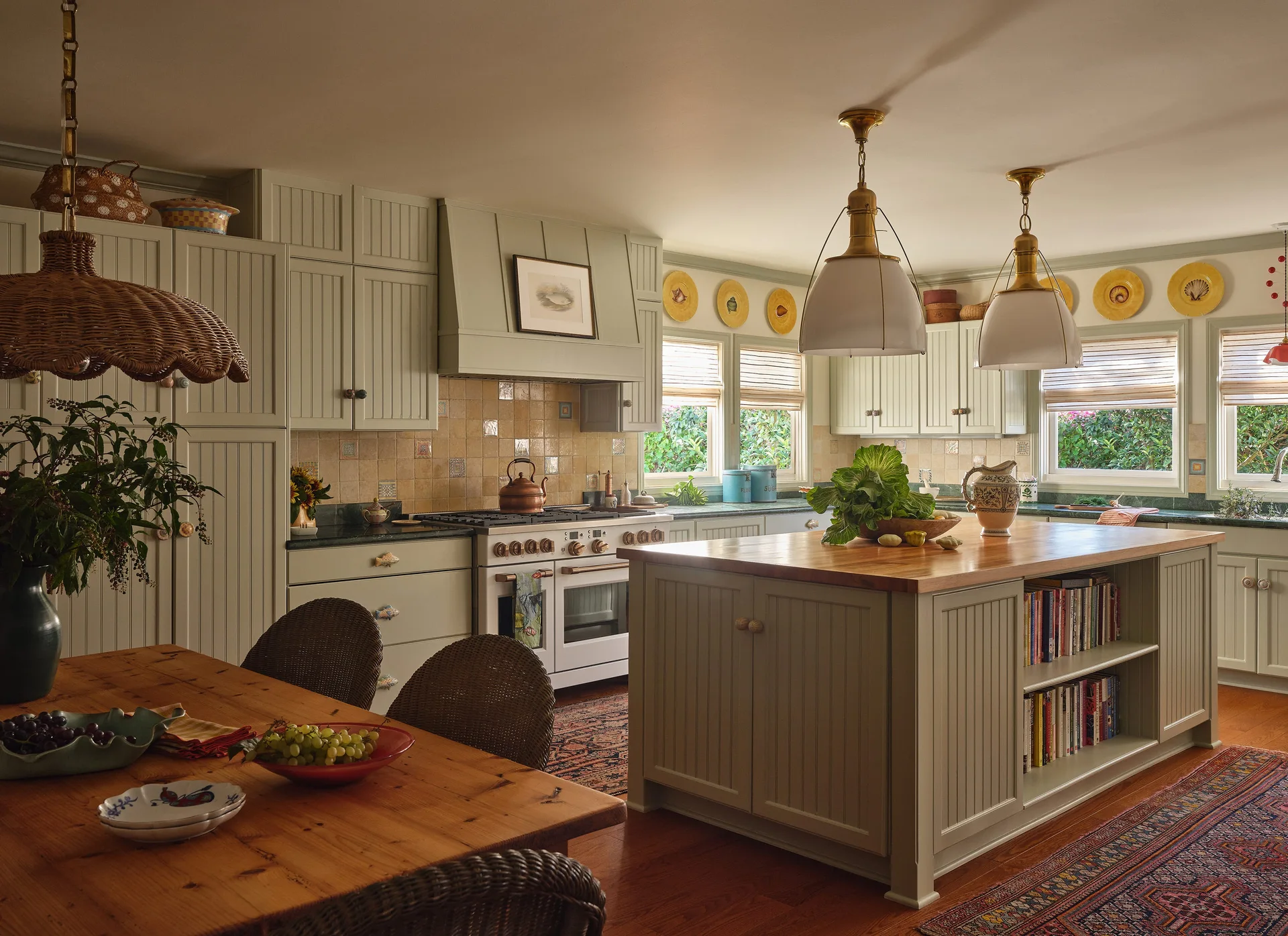 A cottage kitchen features sage green beadboard cabinets, a wood-topped island with bookshelves, and a wooden dining table with wicker chairs.