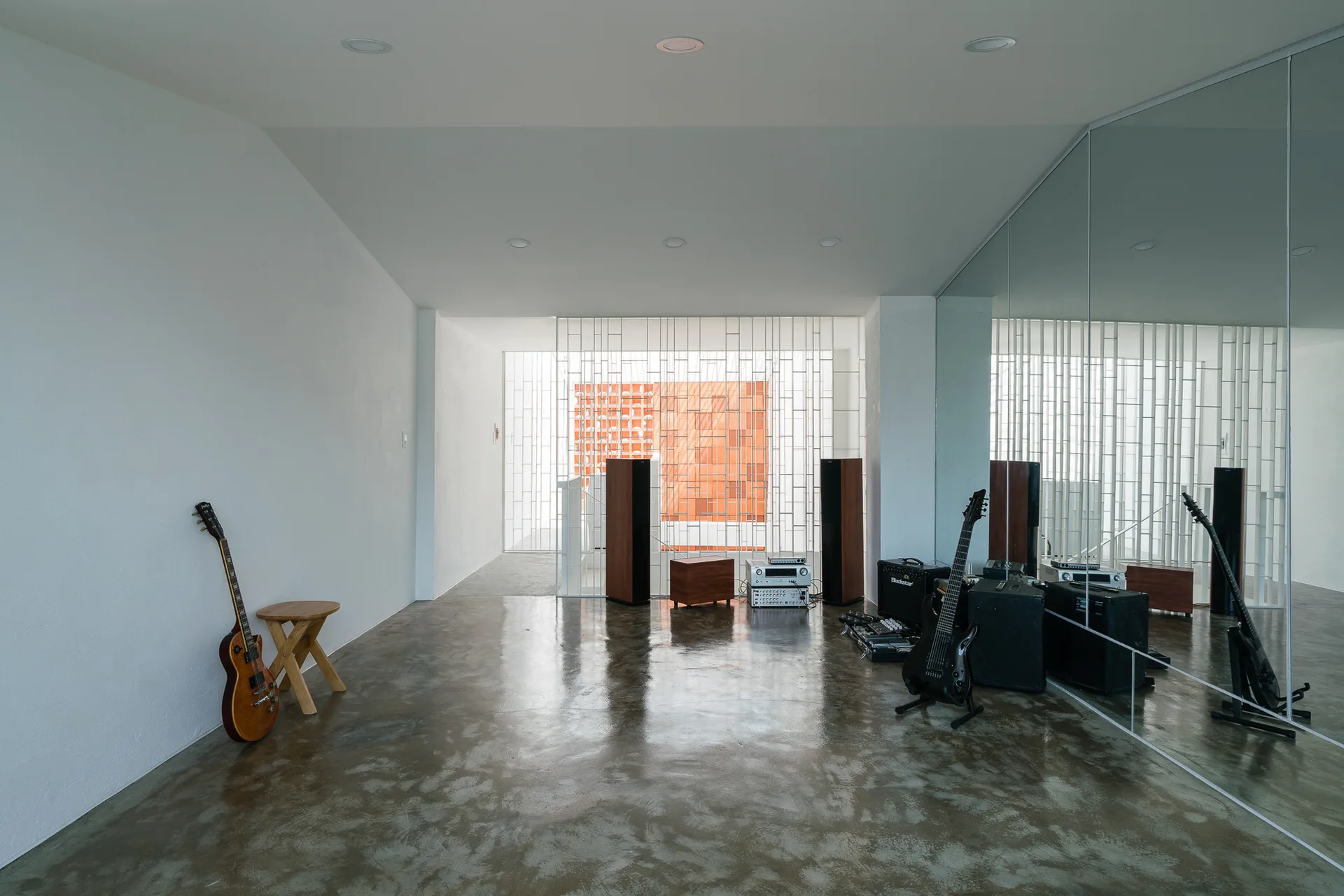 A minimalist music studio features a polished concrete floor, white walls, a large mirrored wall, and audio equipment with guitars.