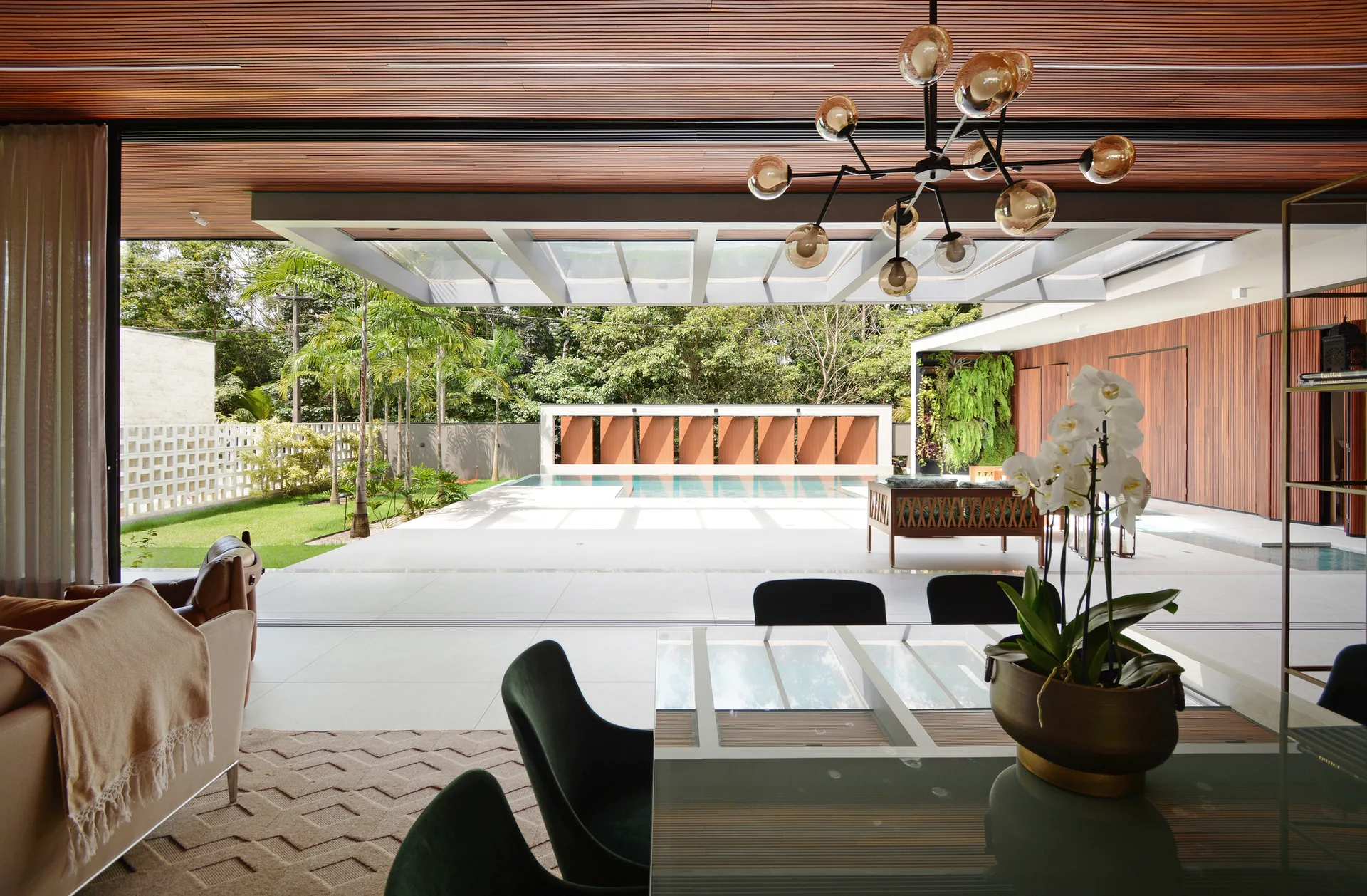 A modern dining room with a glass table and dark chairs opens completely to an outdoor patio with a swimming pool and a wood slat ceiling.