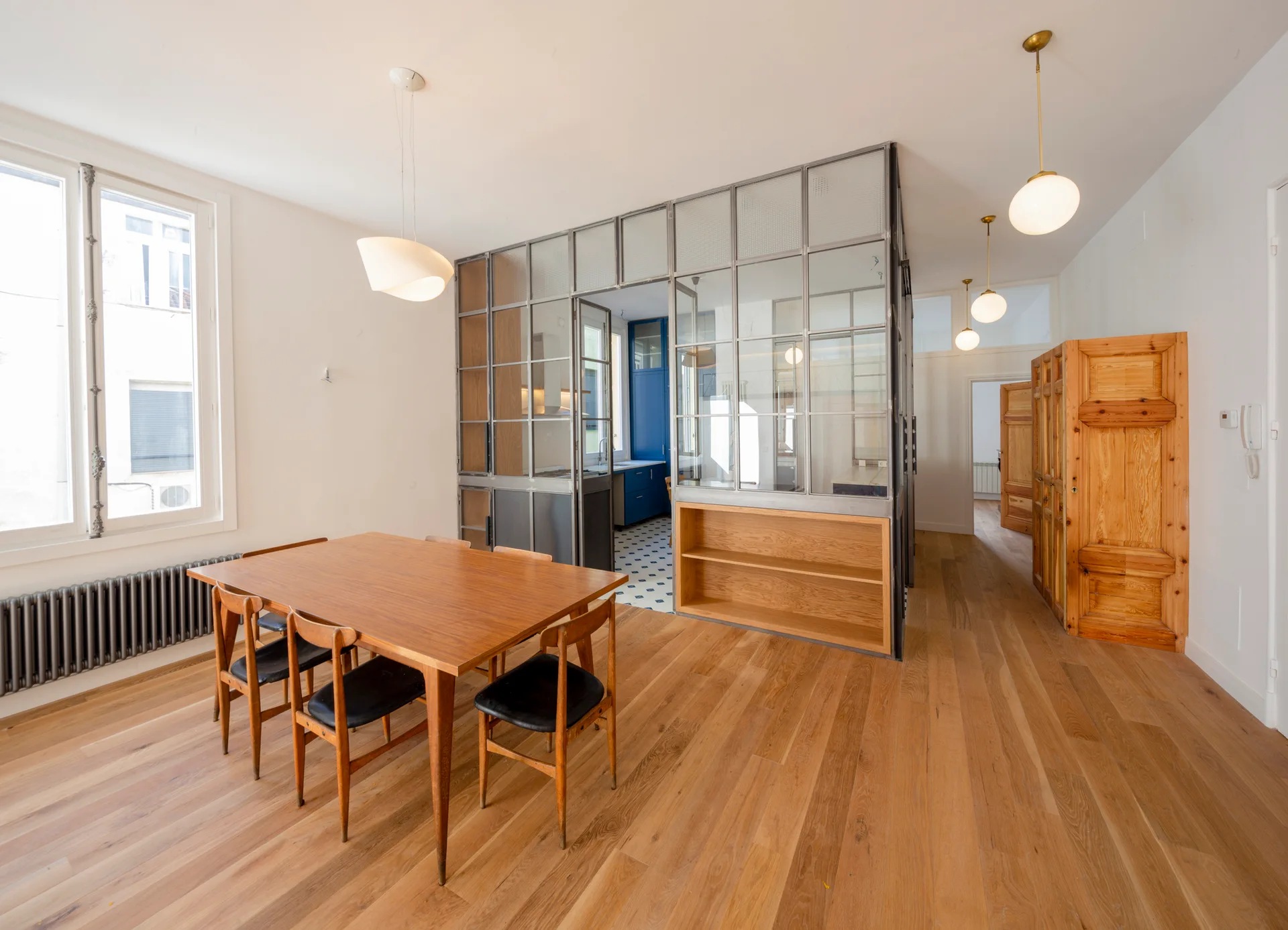 A dining room features a wooden table and chairs next to a glass and metal partition wall that separates the space from a kitchen.