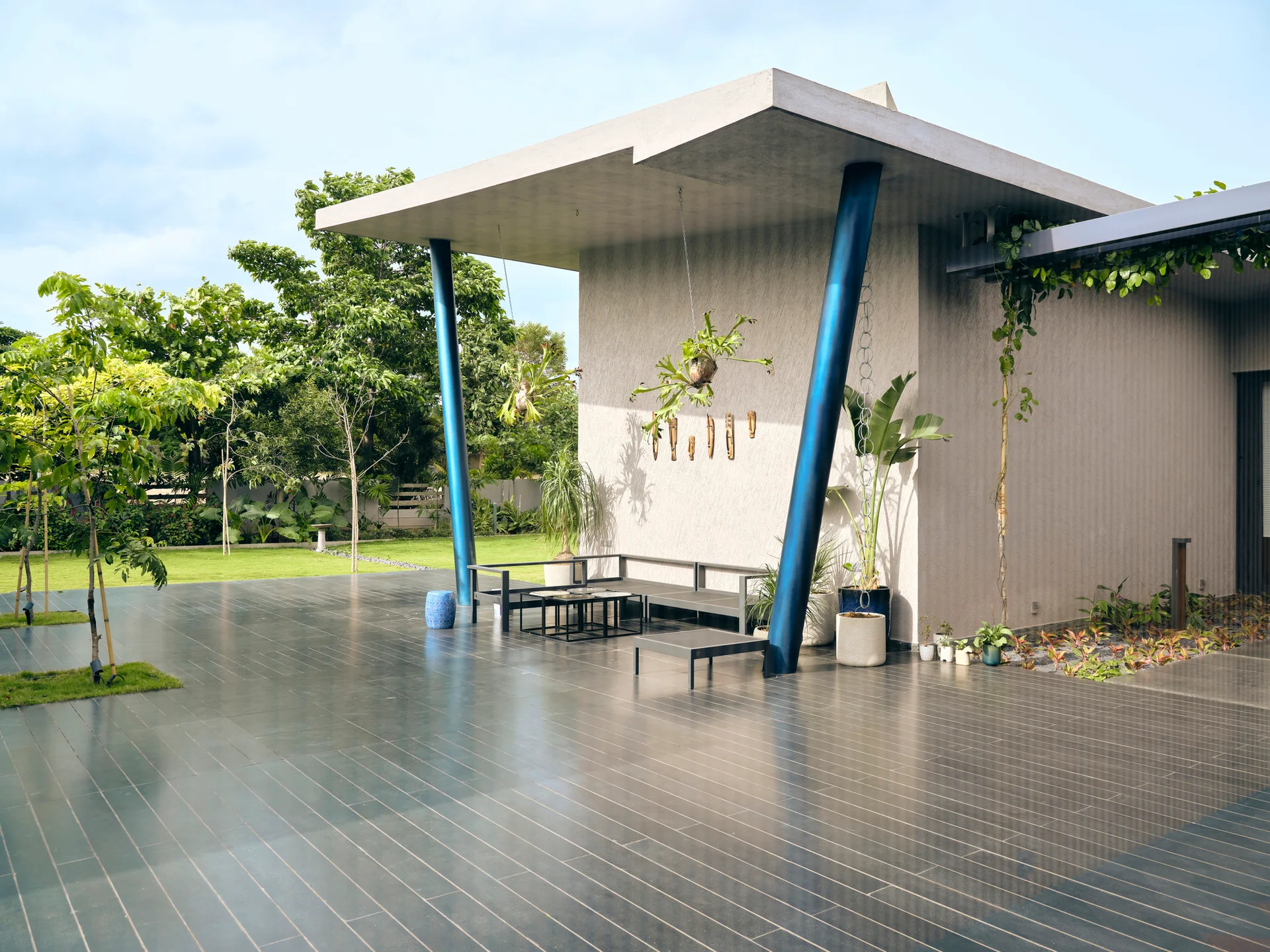 A spacious modern outdoor patio features a concrete canopy supported by metallic blue columns, dark floor tiles, and black metal outdoor furniture.