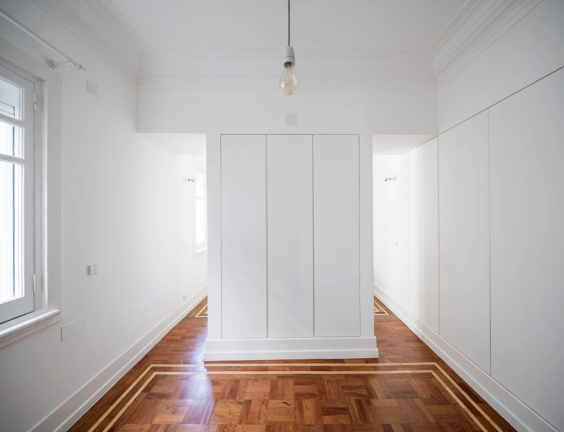 A minimalist room featuring a central white wardrobe partition, detailed wood parquet flooring, and a single hanging bulb.