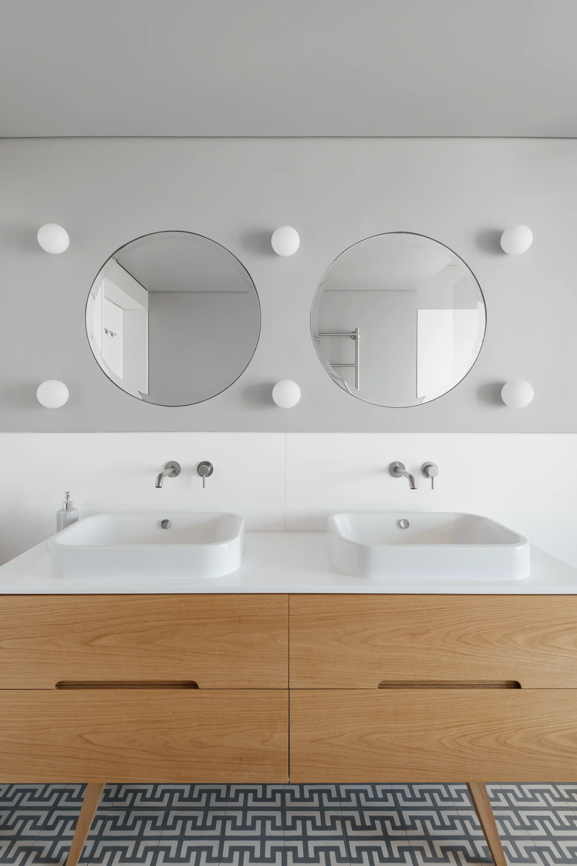 A modern bathroom features a light wood double vanity with two white ceramic vessel sinks, circular mirrors, and grey geometric floor tiles.