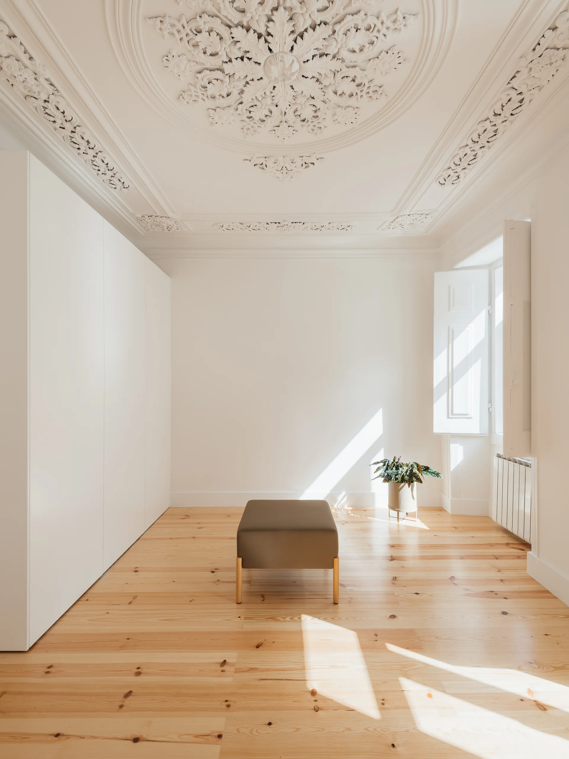 A minimalist white room with a historic ornate plaster ceiling medallion, light wood floors, and a single brown central ottoman.