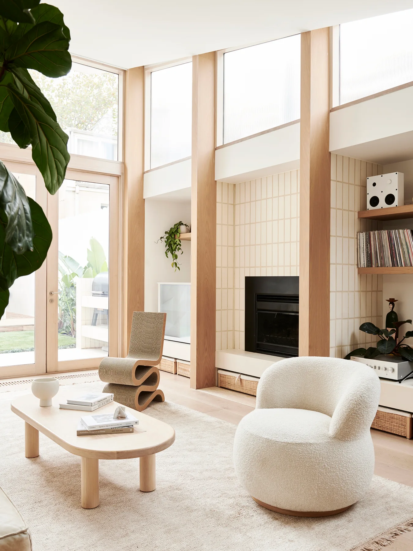 A light-filled living room featuring a grid-tiled fireplace, a cream bouclé armchair, a wooden coffee table, and tall wood-framed windows.