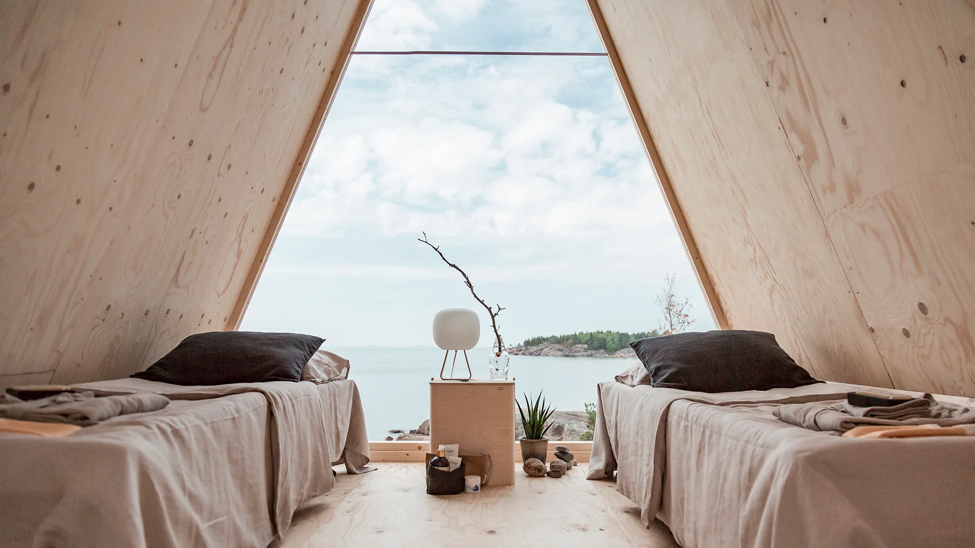 Two twin beds with linen sheets face a large triangular A-frame window overlooking the sea inside a minimalist plywood cabin bedroom.