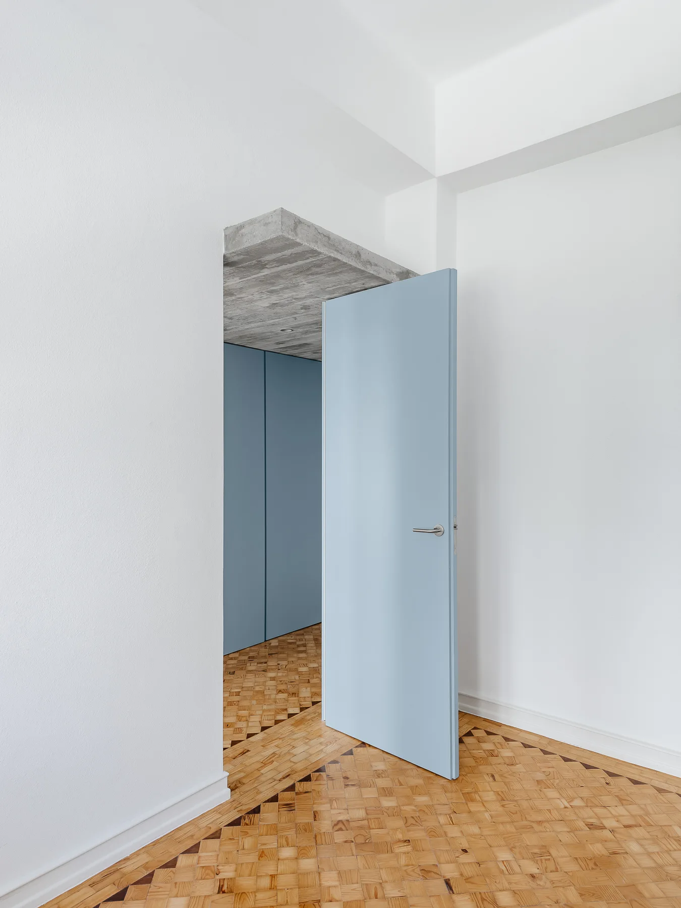 A light blue door stands open in a white room, revealing a hallway with geometric parquet wood floors and an exposed concrete lintel above.