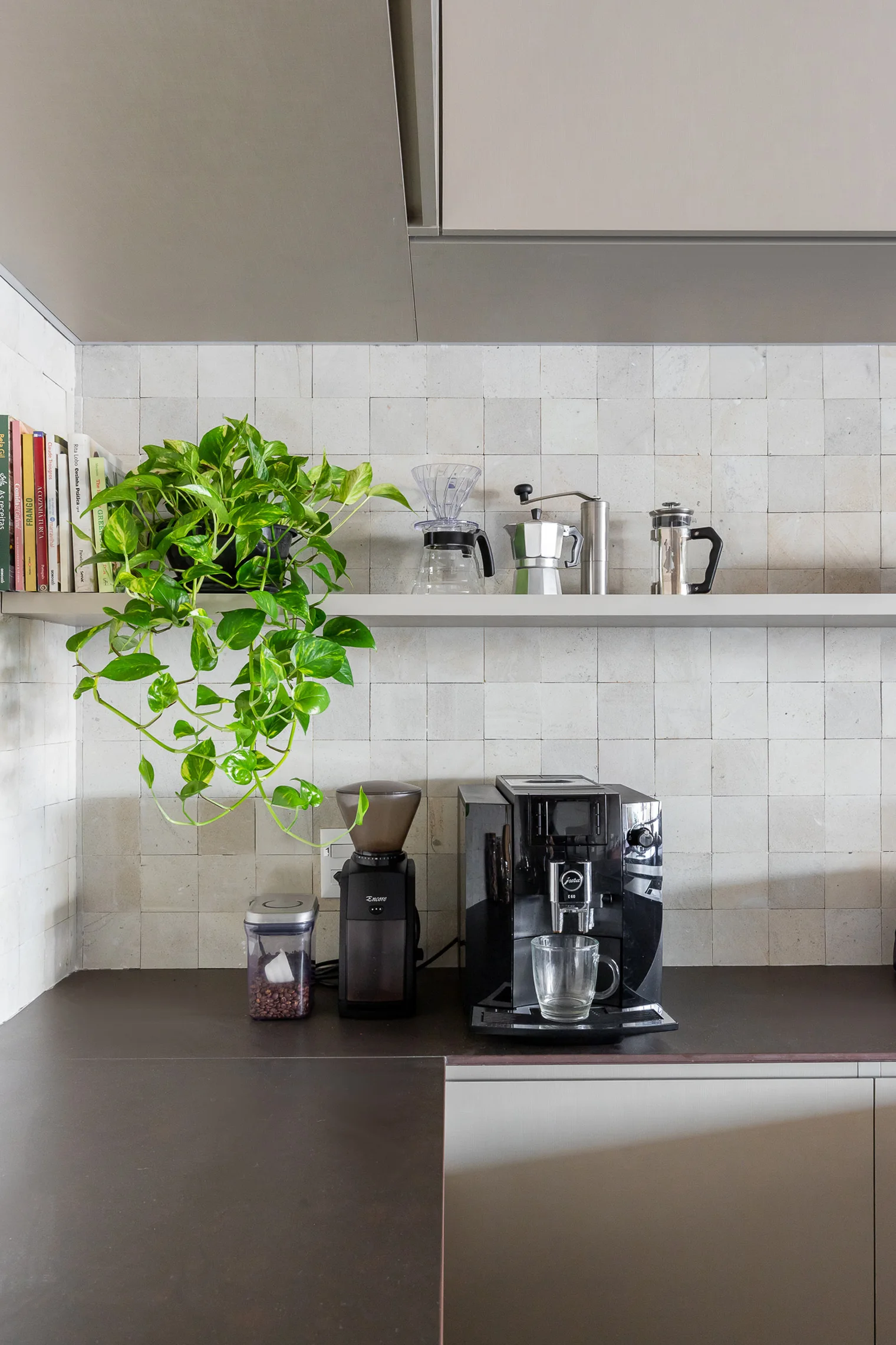A modern kitchen coffee station features a black espresso machine on a dark countertop below a floating shelf with books, plants, and coffee gear.