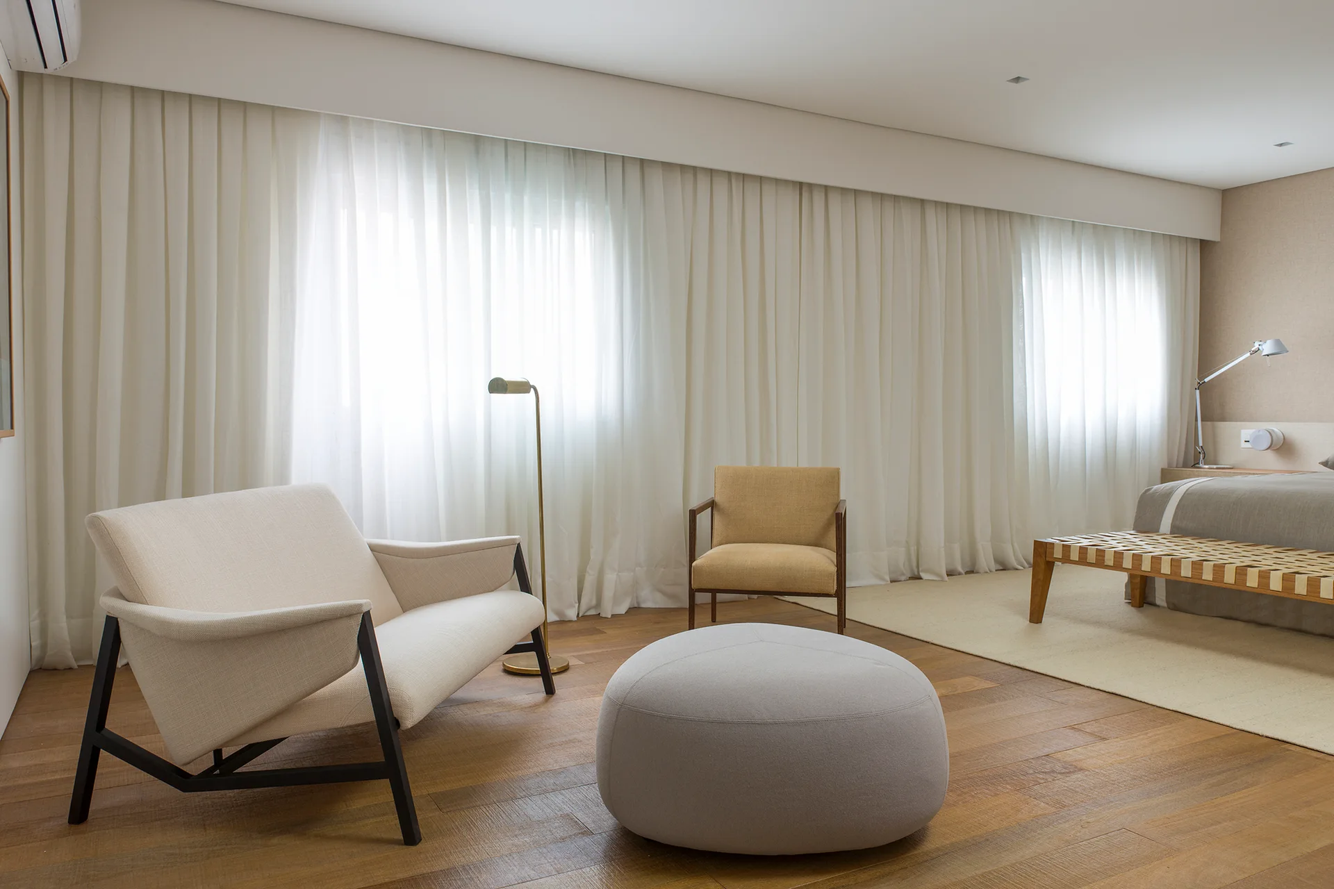 A minimalist bedroom features a cream armchair, a round grey ottoman, and a mustard yellow chair in front of floor-to-ceiling sheer white curtains.