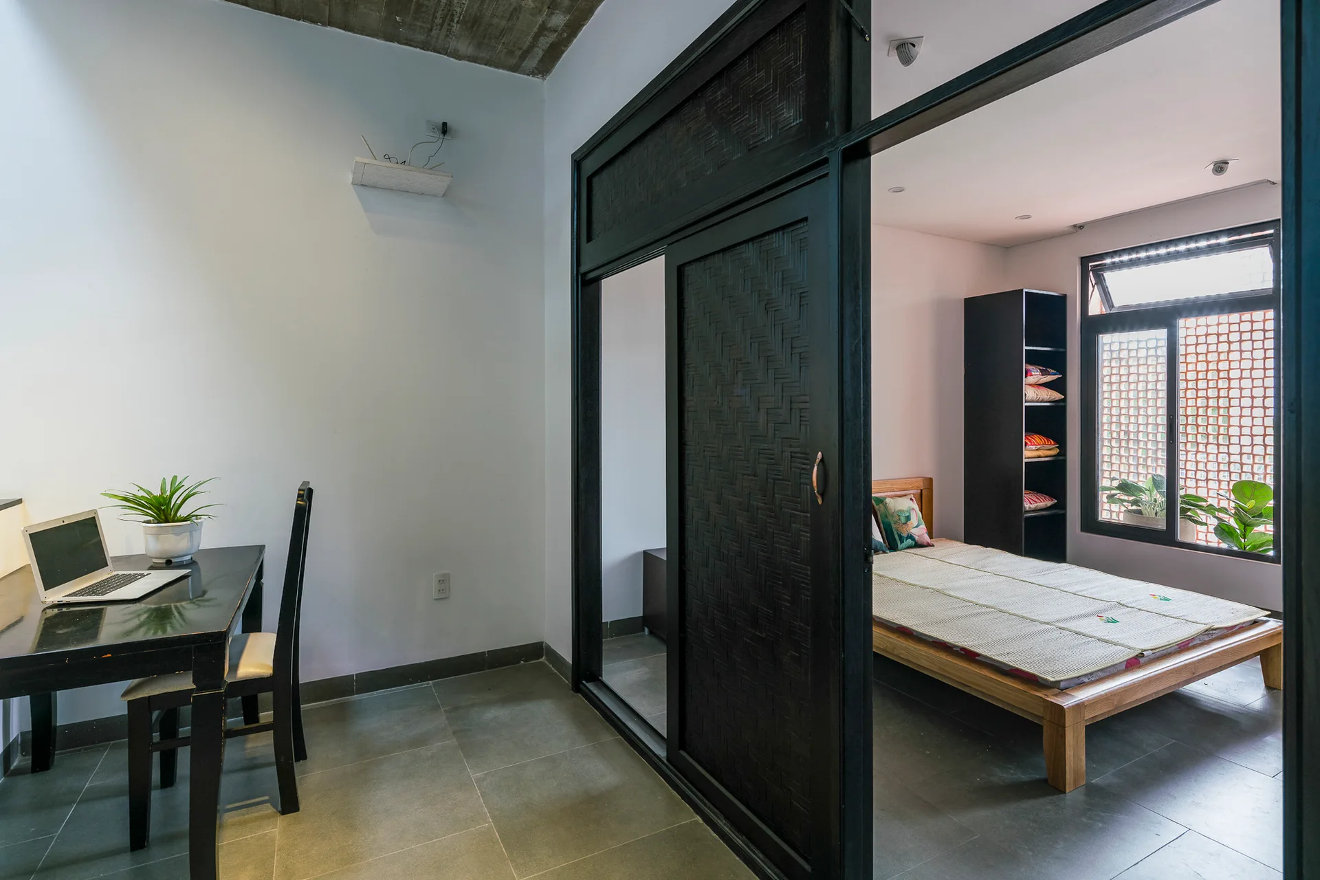 A minimalist workspace with a black desk sits adjacent to a bedroom featuring a wooden bed frame, separated by a black woven sliding door.