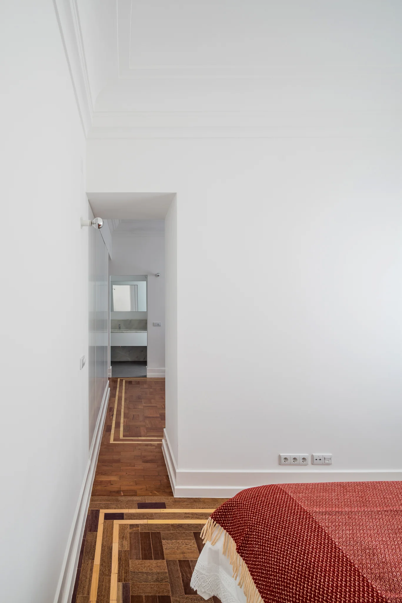 A white bedroom with detailed parquet wood floors features a doorway opening to a bathroom with a marble-topped vanity.