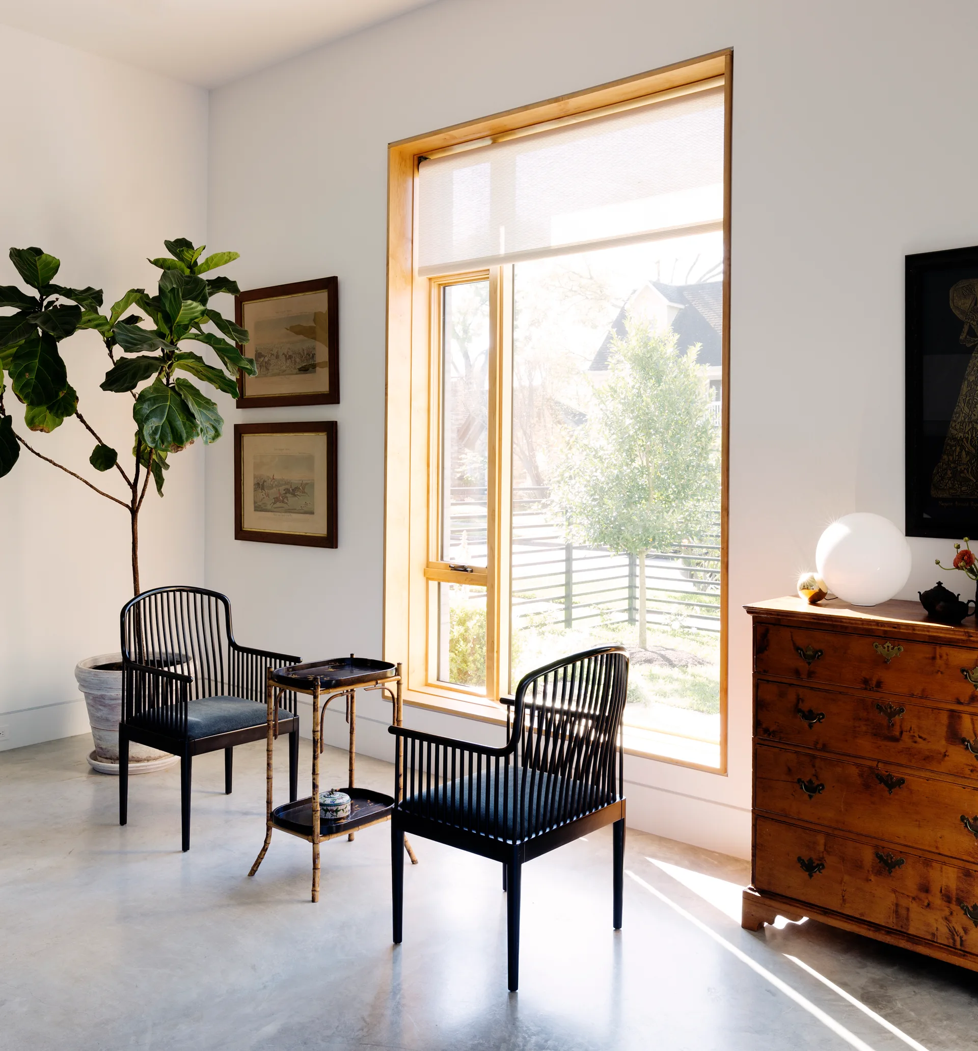 Two black spindle-back chairs face each other next to a bamboo side table in front of a large wood-framed window with a polished concrete floor.