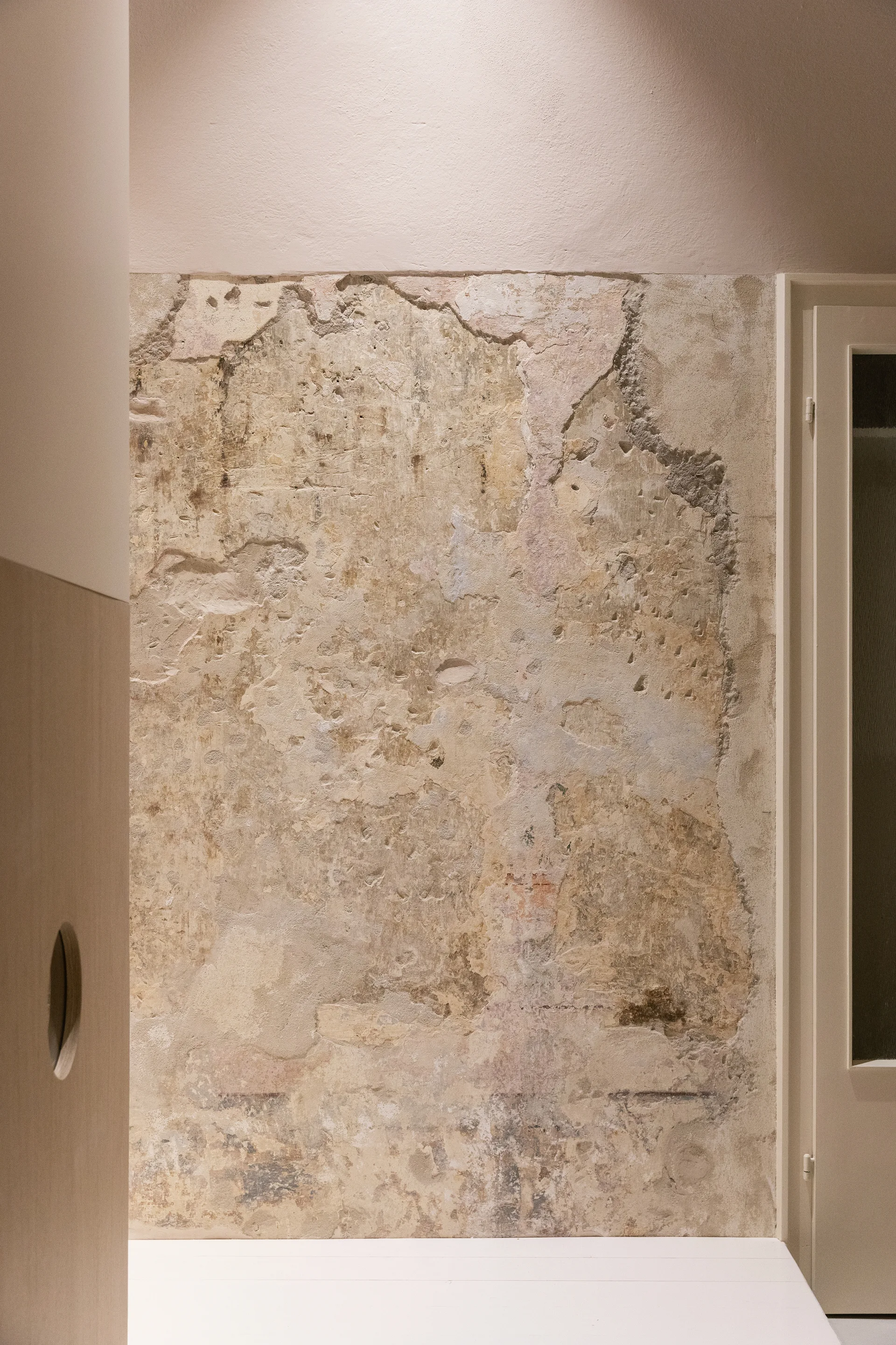 An exposed weathered plaster wall in neutral beige tones is framed by smooth modern wood paneling and a clean white doorway.