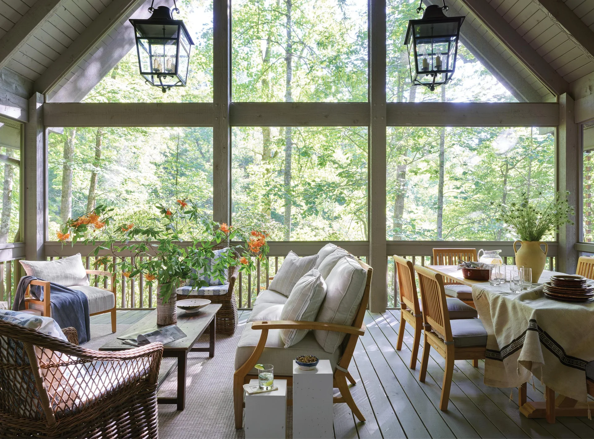 A spacious screened-in porch featuring a wood dining set, cushioned sofa, wicker chairs, and two hanging black lanterns overlooking a forest.