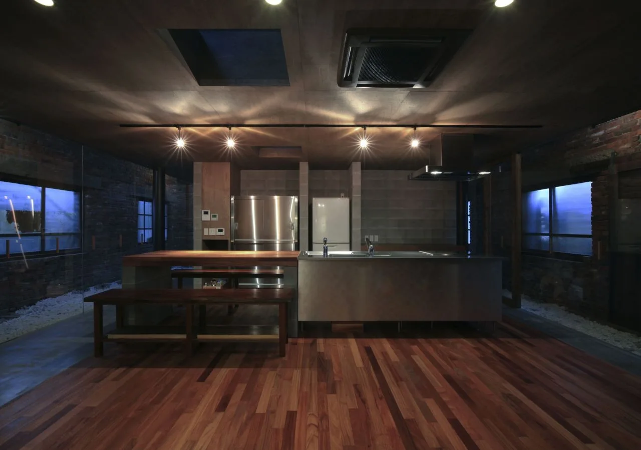 An industrial kitchen features a central stainless steel and wood island with wooden bench seating, exposed brick walls, and a dark wood ceiling.