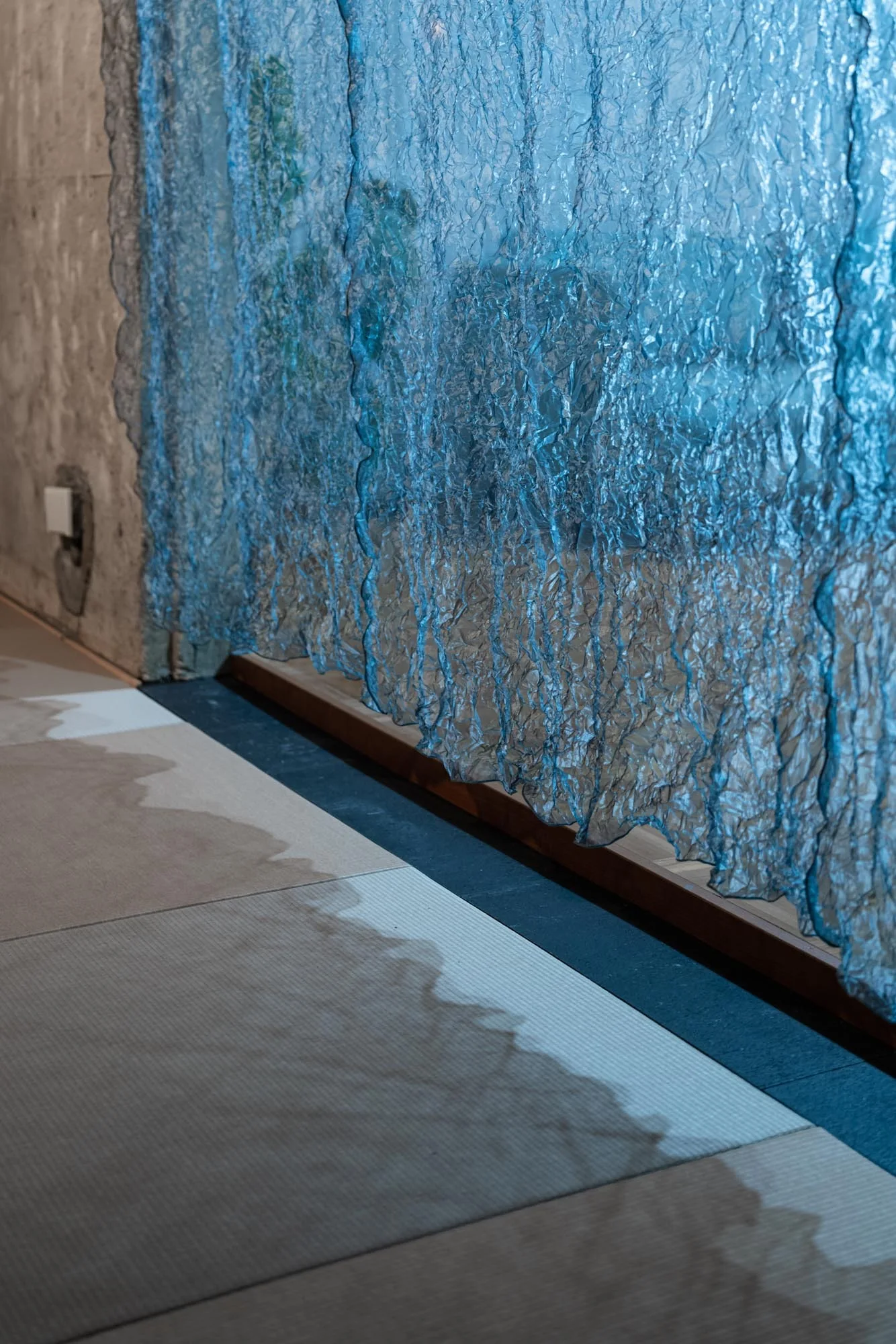A translucent crinkled blue curtain hangs next to a textured concrete wall, casting soft blue shadows onto a light-colored floor.