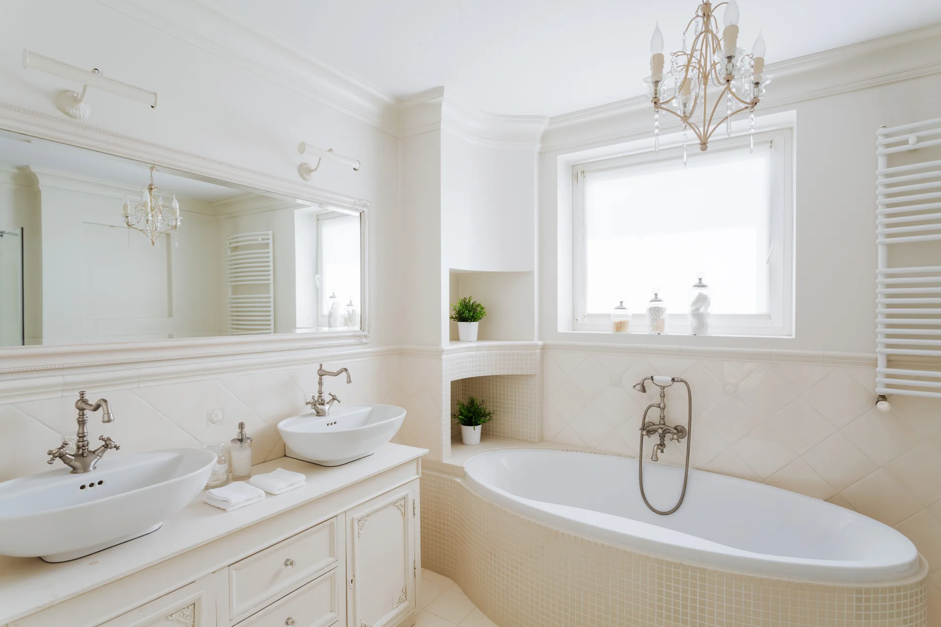 A cream bathroom featuring a double vanity with vessel sinks, a tiled oval bathtub under a window, and a built-in recessed corner niche.
