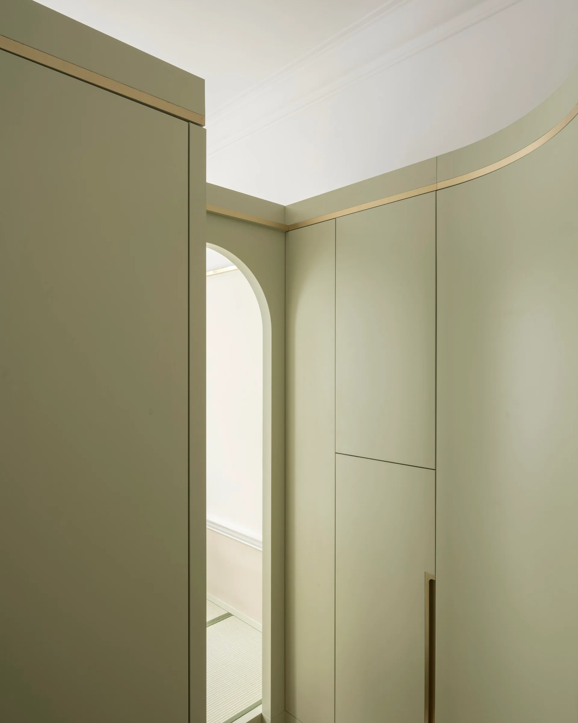 Matte sage green built-in cabinetry featuring a gold metal inlay trim along a curved wall next to an arched doorway.