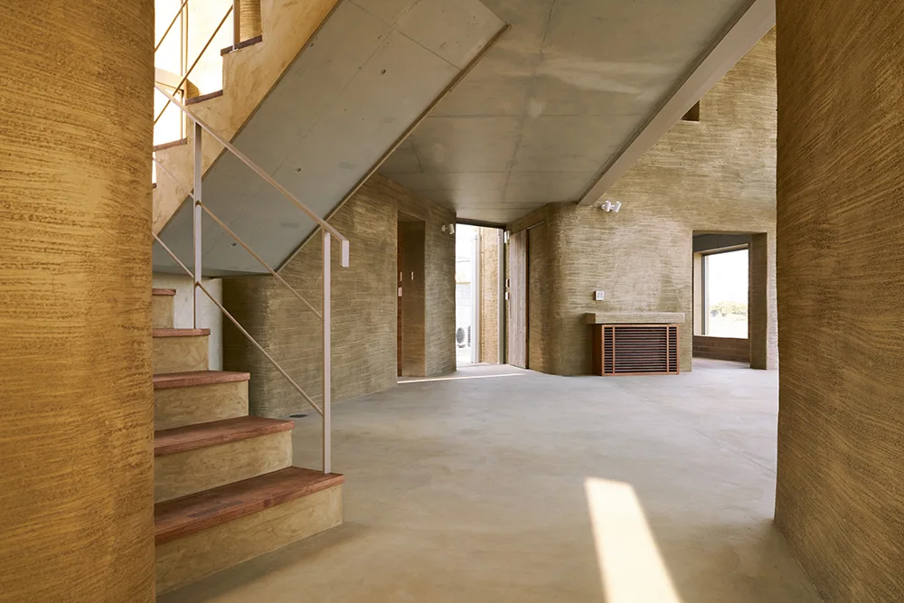 An architectural entryway features highly textured, curved earthen walls, a concrete floor, and a staircase with wooden treads and a slim metal railing.
