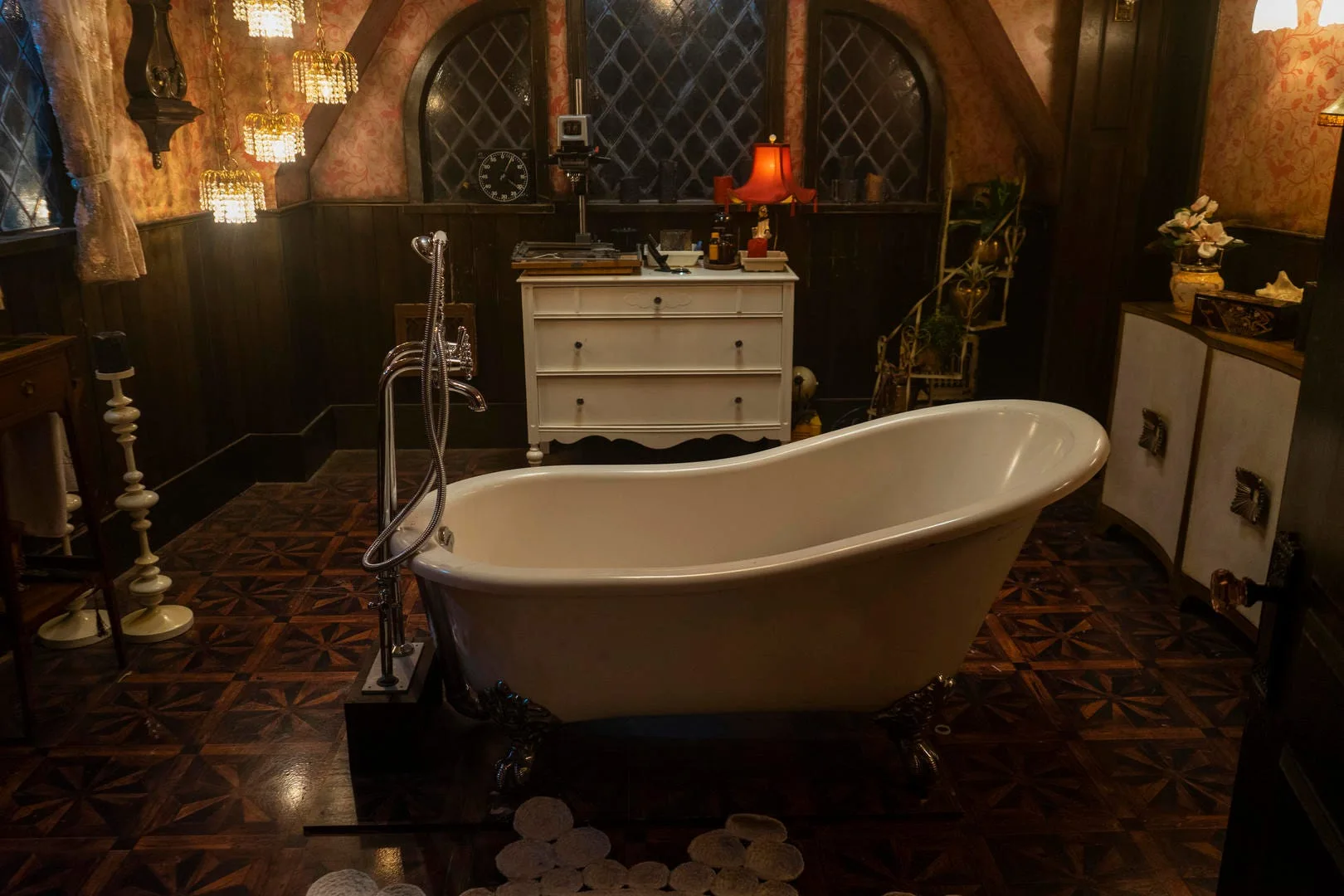 A vintage bathroom features a white clawfoot tub, dark wood wainscoting, floral wallpaper, arched lattice windows, and a white dresser.