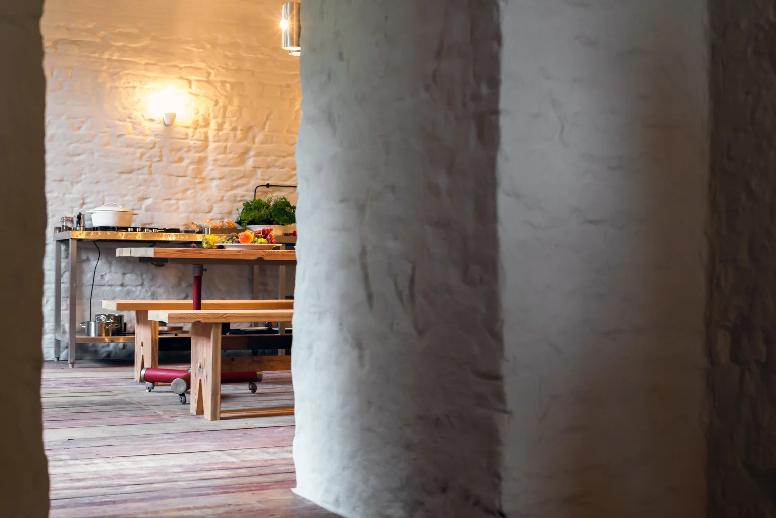 A rustic kitchen viewed through an archway, featuring a wooden dining table and benches in front of a textured white brick wall.
