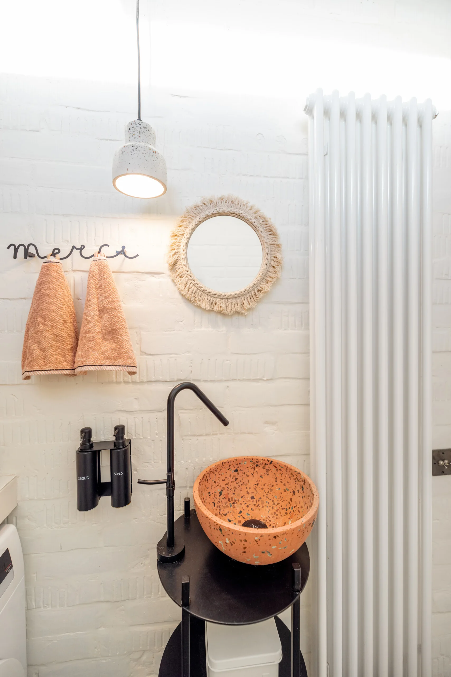 A peach terrazzo vessel sink sits on a black stand in front of a white brick wall with a fringed round mirror and pendant light.
