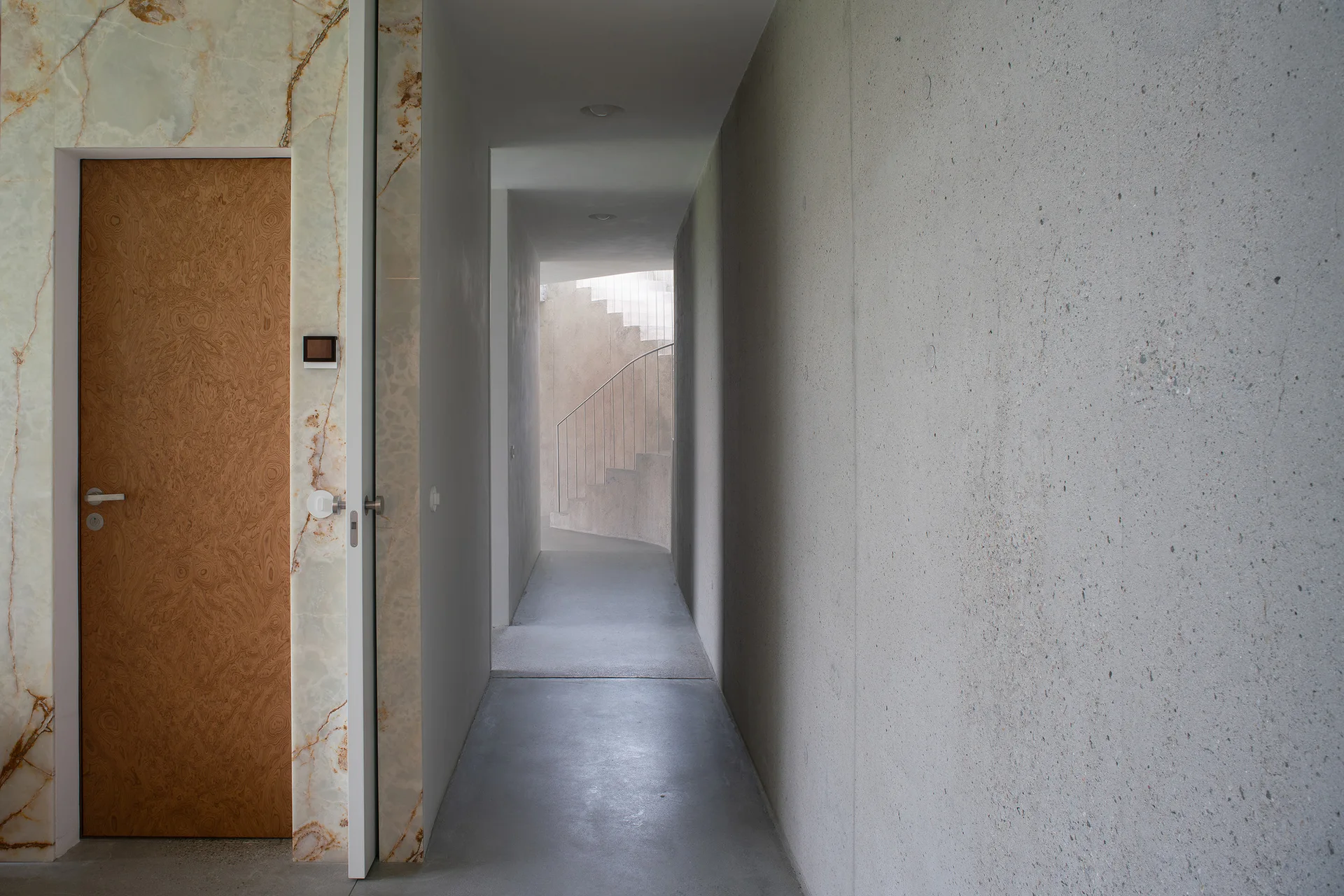 A long minimalist concrete hallway features a wood door framed in veined stone cladding leading to a curved staircase in the background.