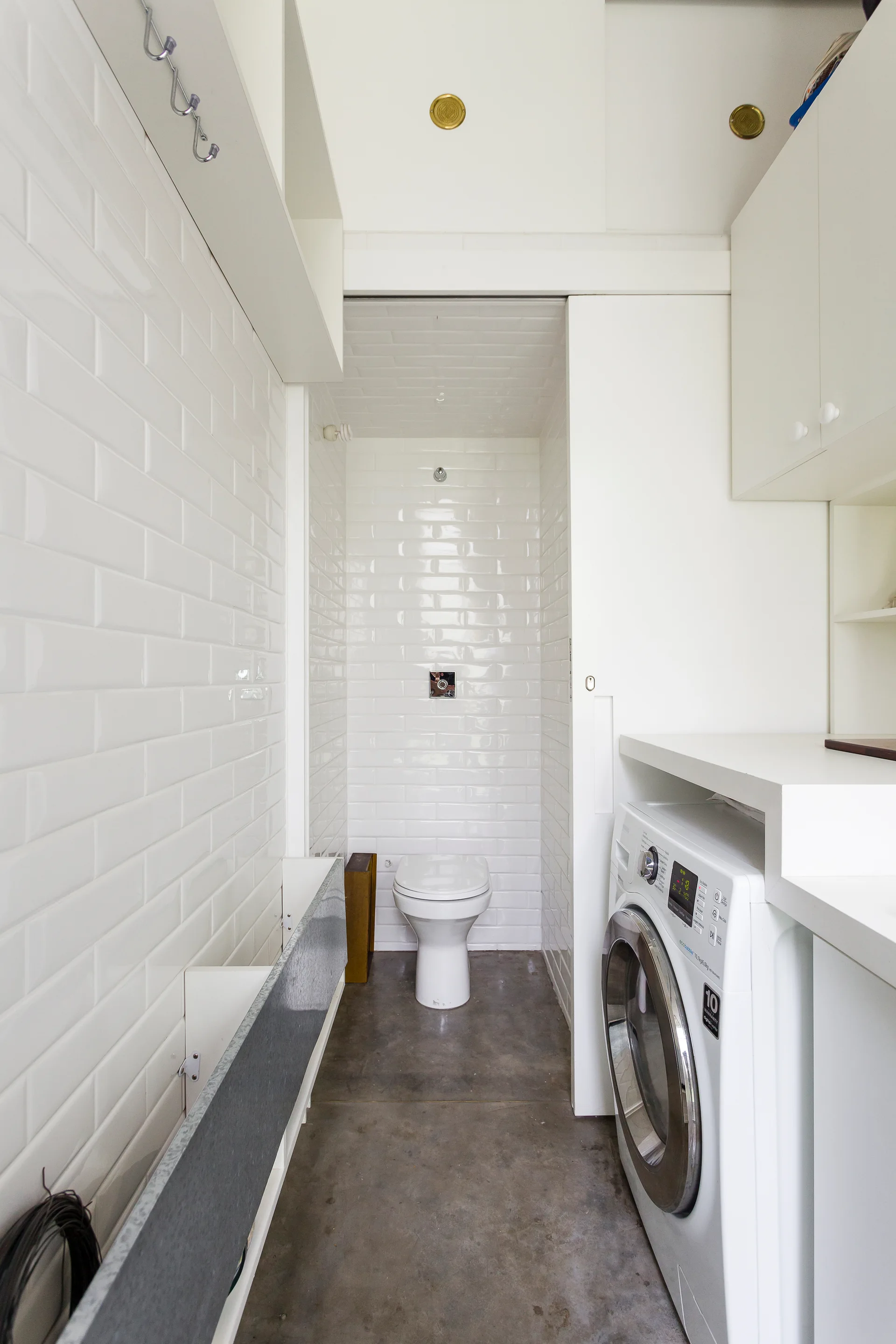 A narrow bathroom laundry room combo featuring white subway tiled walls, a toilet at the end, and a front-load washing machine under white cabinetry.