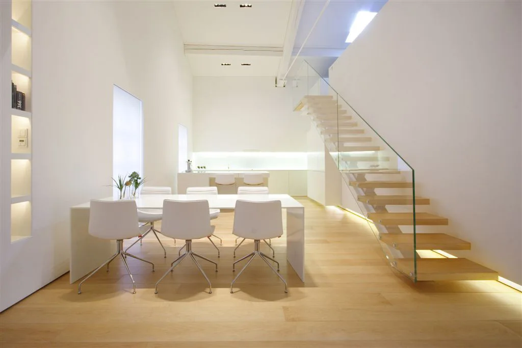 A minimalist white dining room featuring a dining table and chairs adjacent to a floating wooden staircase with a transparent glass railing.