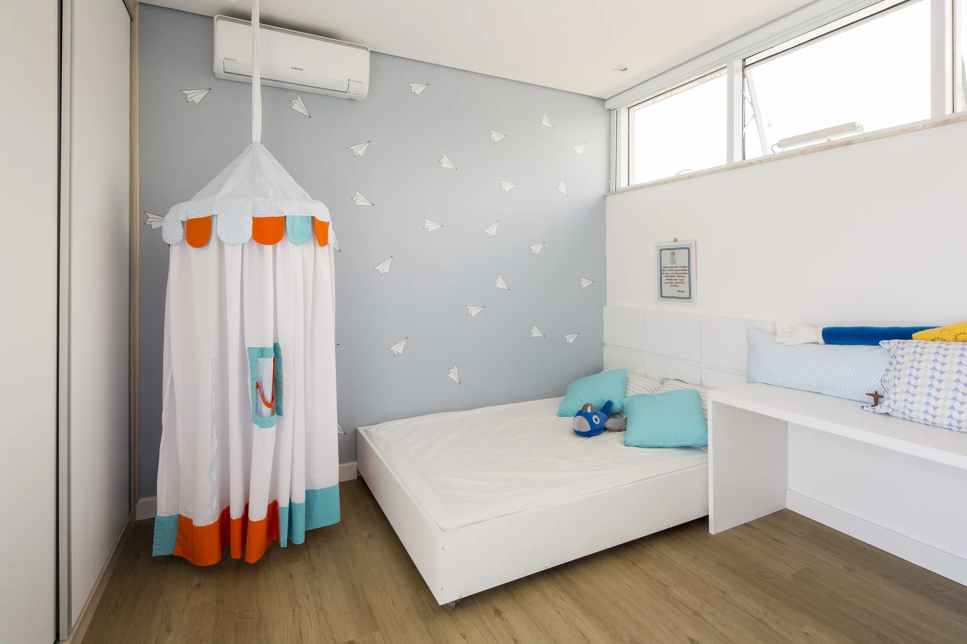 A kids bedroom features a white platform bed, blue accent pillows, a hanging canopy tent, and a light blue wall decorated with paper plane decals.