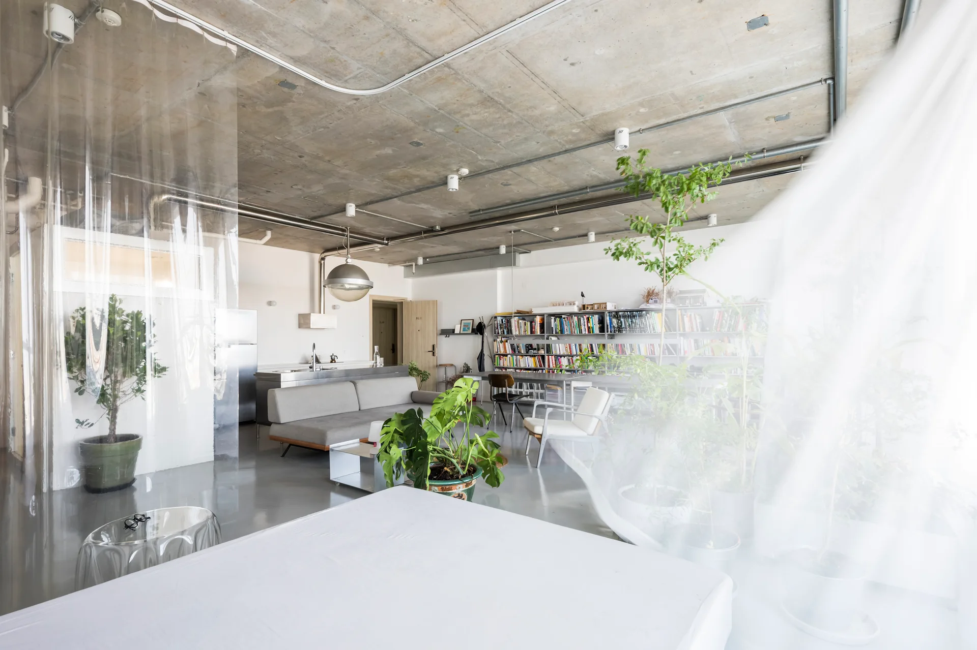 An industrial loft living room with an exposed concrete ceiling, a grey sofa, a large metal bookshelf, and potted plants.