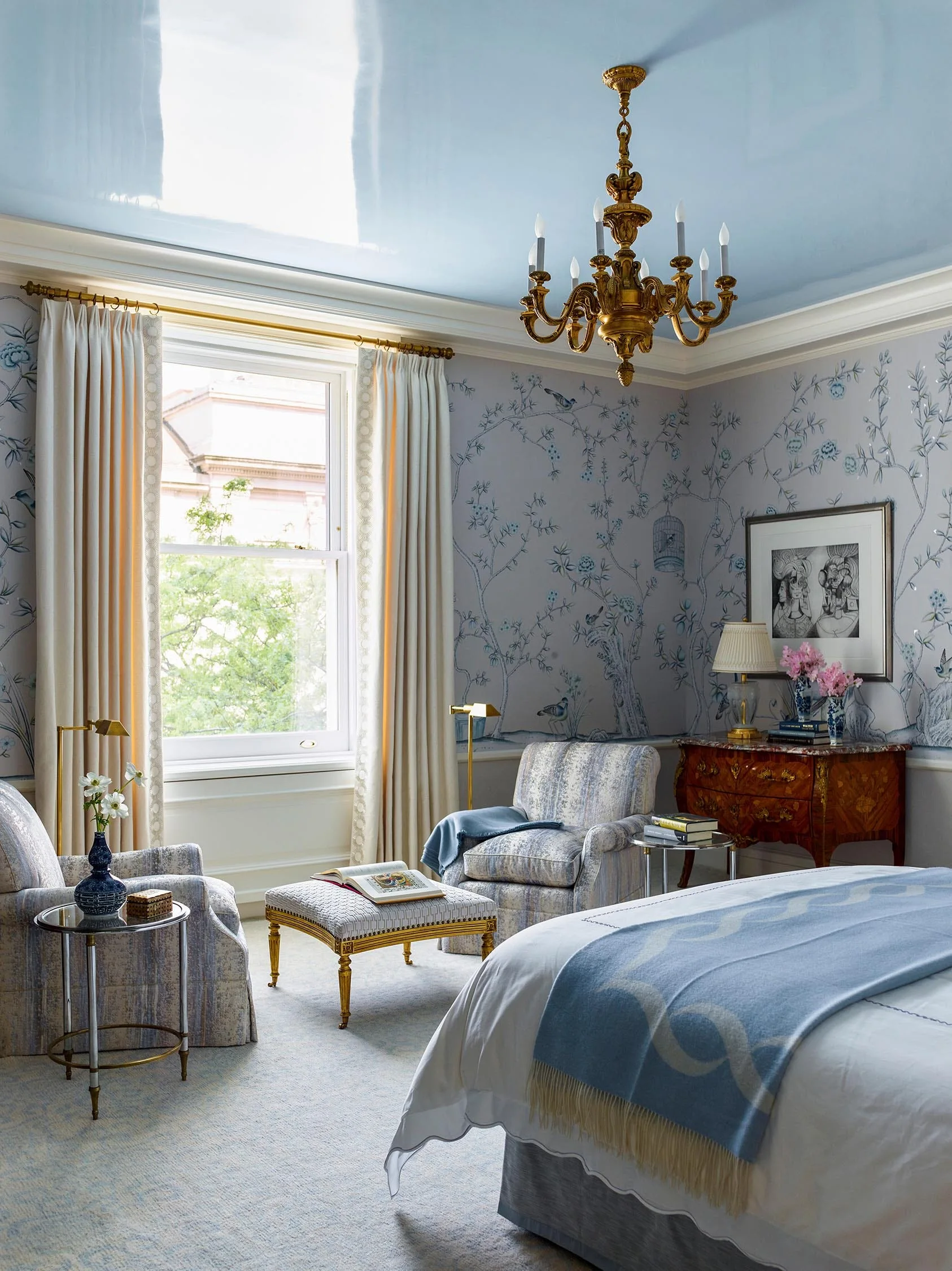 A traditional bedroom features a light blue lacquered ceiling, patterned chinoiserie wallpaper, two upholstered armchairs, and an ornate gold chandelier.