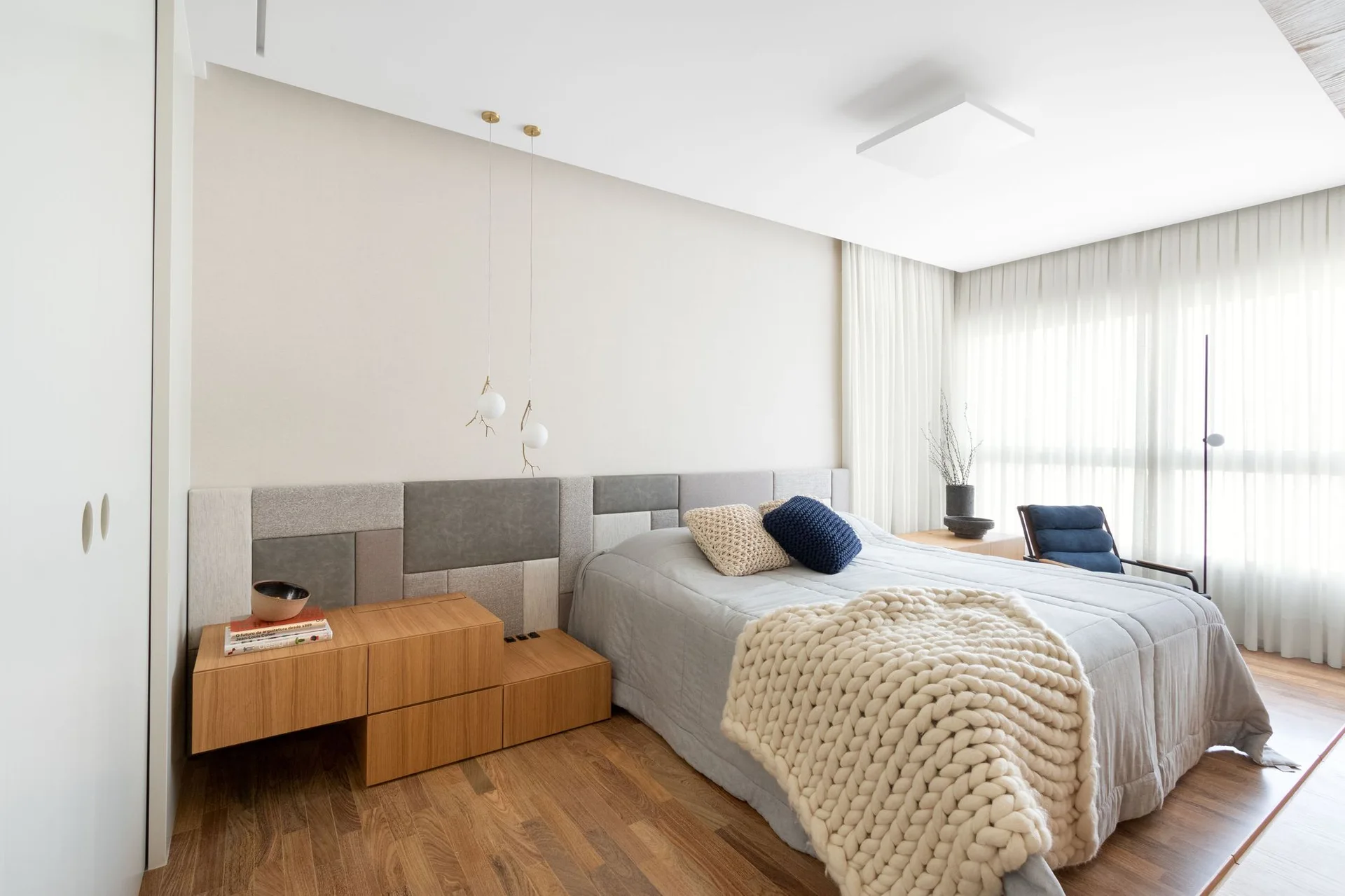 A modern bedroom features a segmented fabric headboard, a low wooden nightstand with books, and a bed draped in a chunky knit throw.