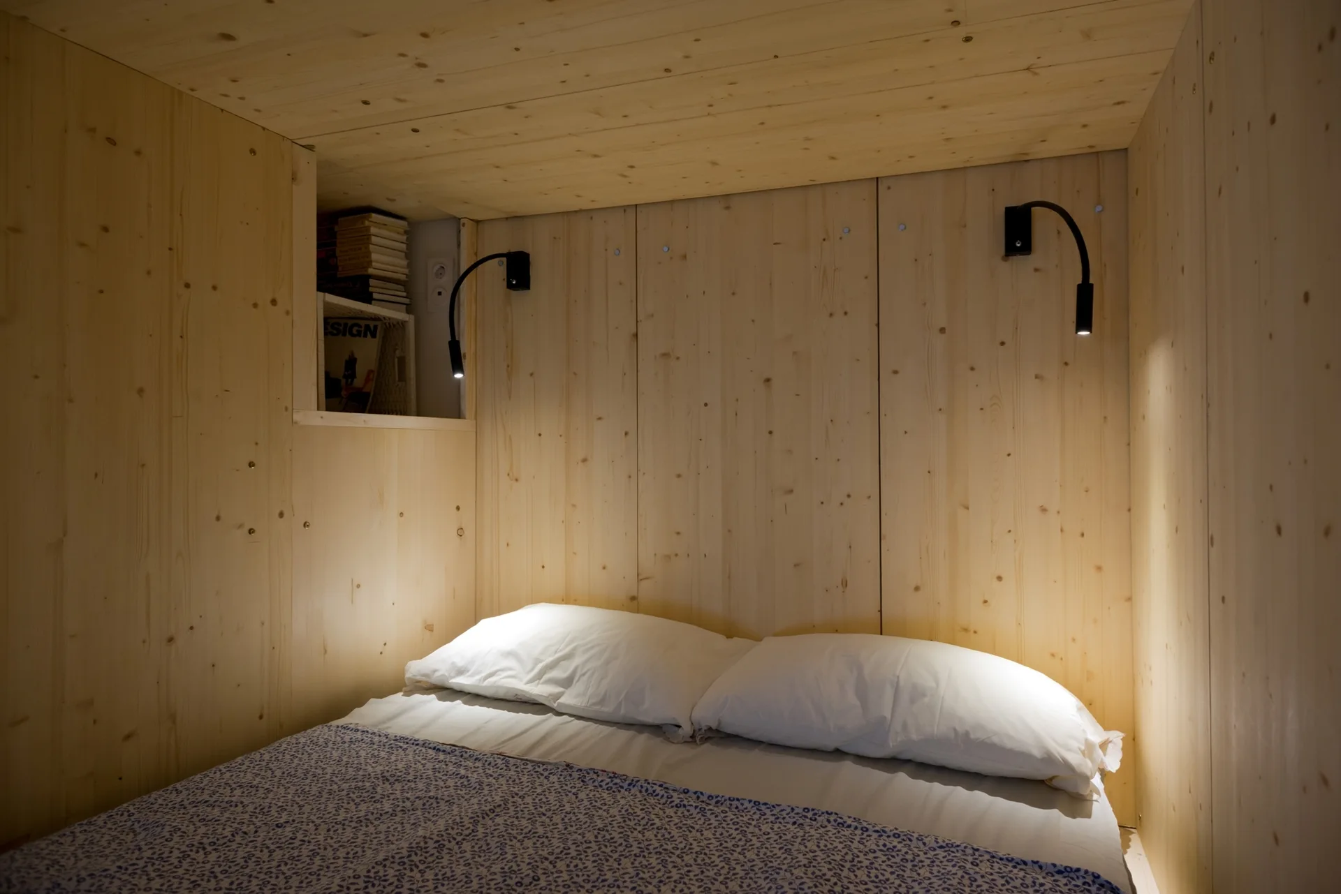 A bed with white pillows and a blue patterned cover is tucked into an alcove with light pine wood paneled walls, ceiling, and built-in shelving.