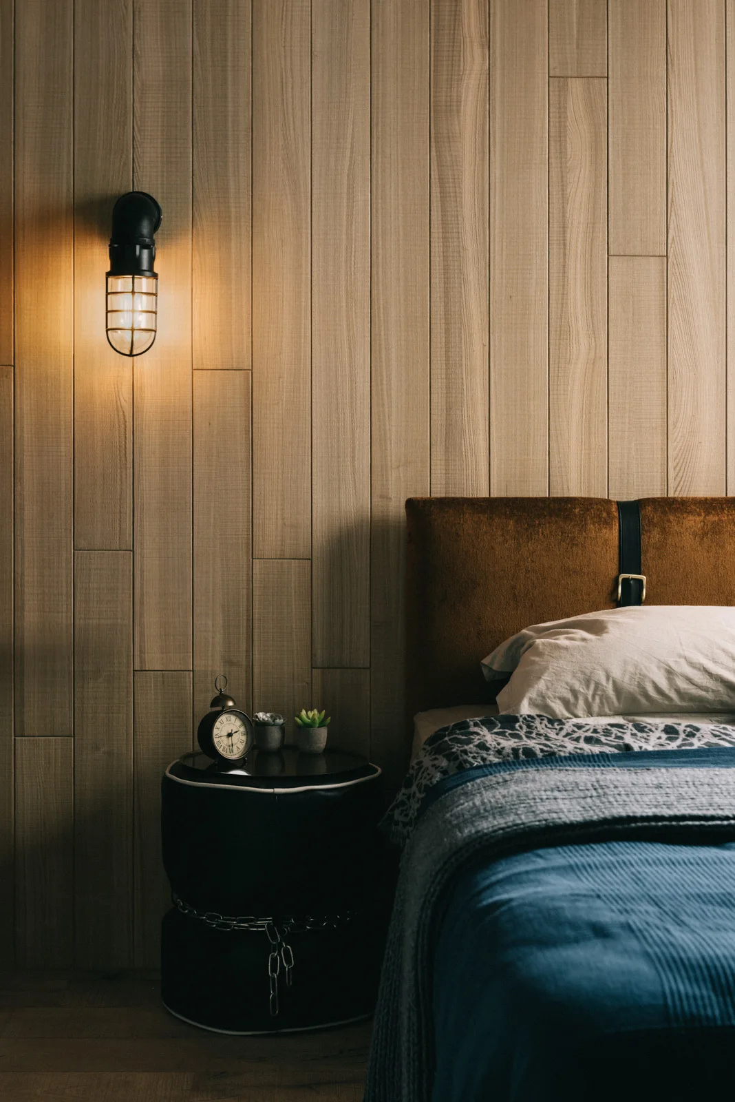 A bedroom features a brown headboard against vertical wood wall paneling, a black circular nightstand, and an illuminated industrial cage wall sconce.