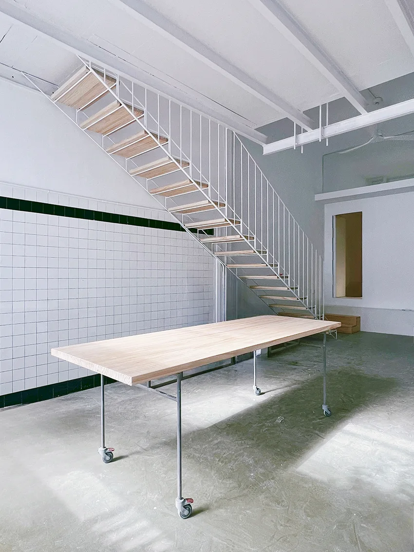 An open interior space featuring a minimalist white metal staircase with wooden treads and a large light-wood table on wheels against a tiled wall.