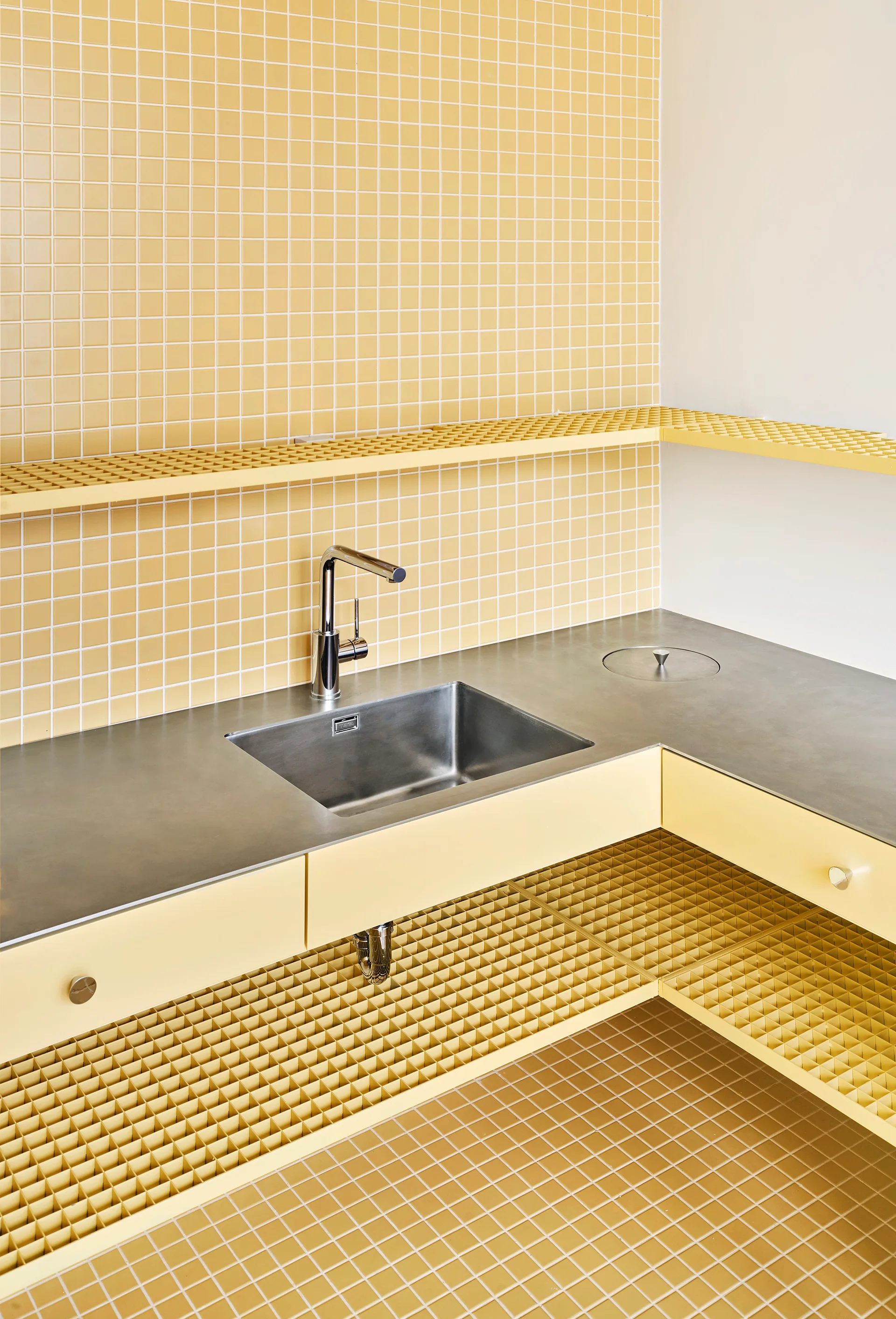 A stainless steel kitchen sink and grey countertop are surrounded by yellow square tile walls and a matching yellow grid-patterned open shelf.