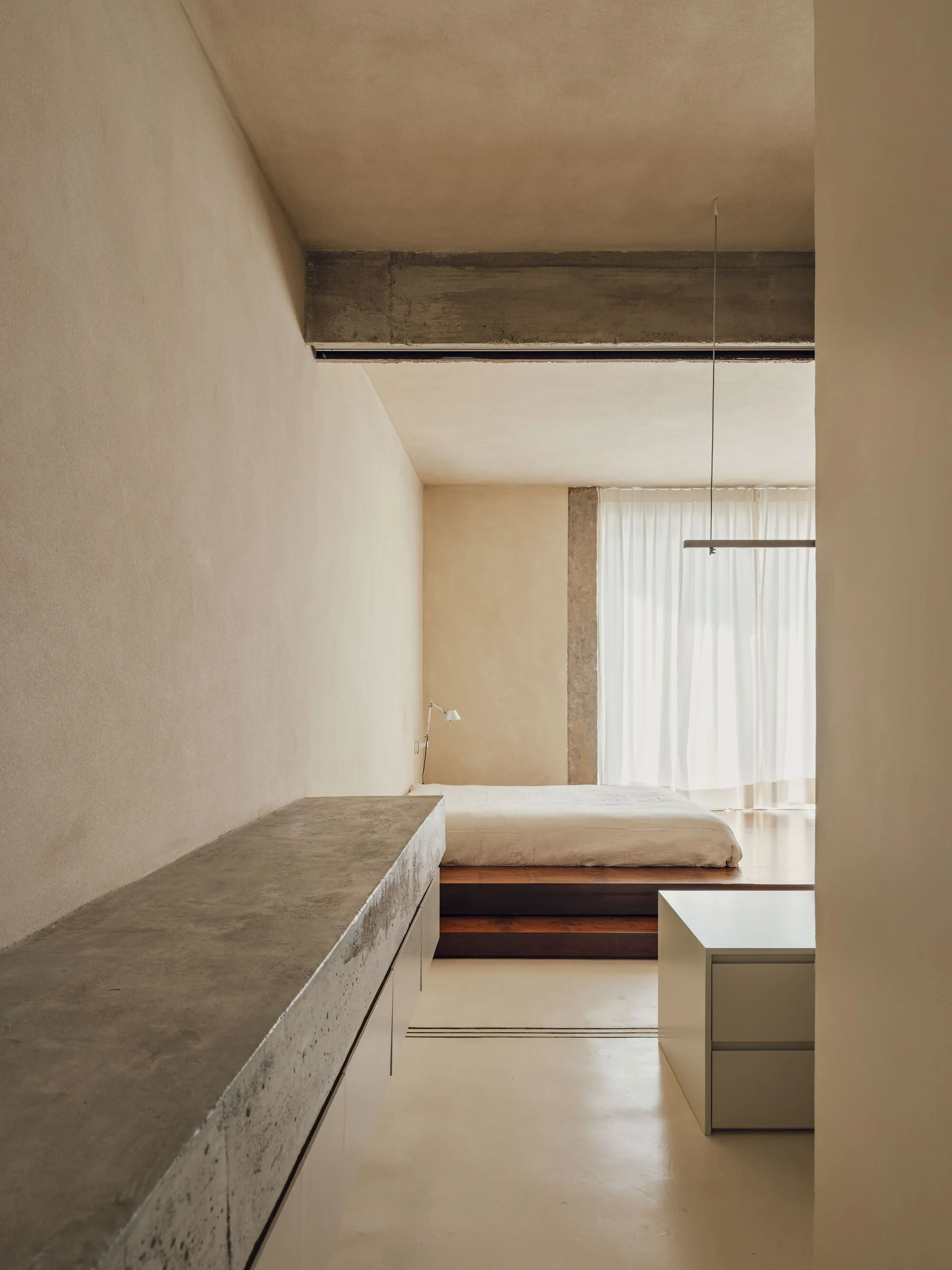 A minimalist bedroom features a low wooden platform bed near a curtained window, viewed past a raw concrete console and textured plaster walls.