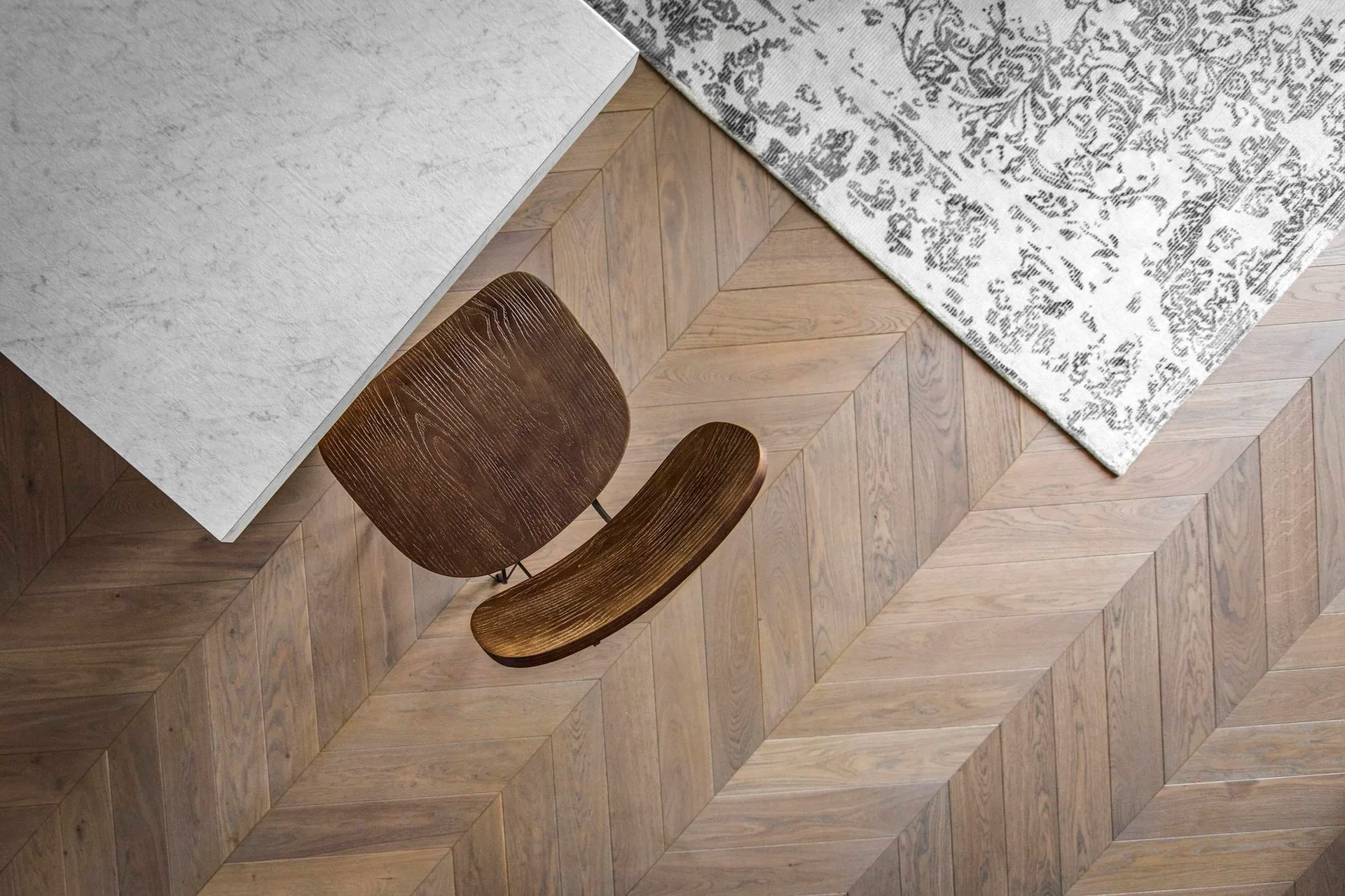 An overhead view of a dark wooden dining chair next to a light grey stone table on herringbone hardwood flooring.