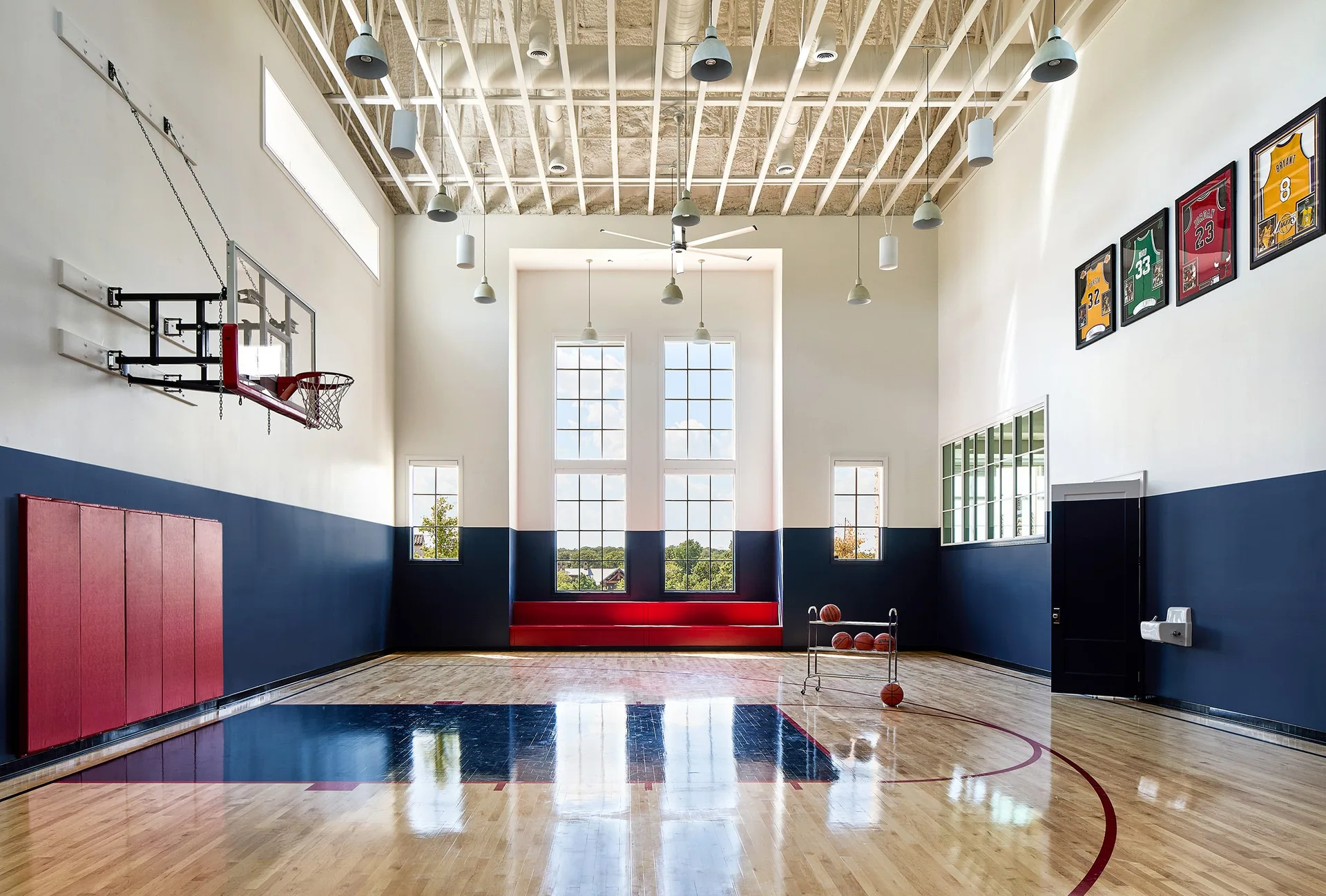 An indoor basketball court features glossy wood floors, navy and white painted walls, a wall-mounted hoop, and a high exposed grid ceiling.