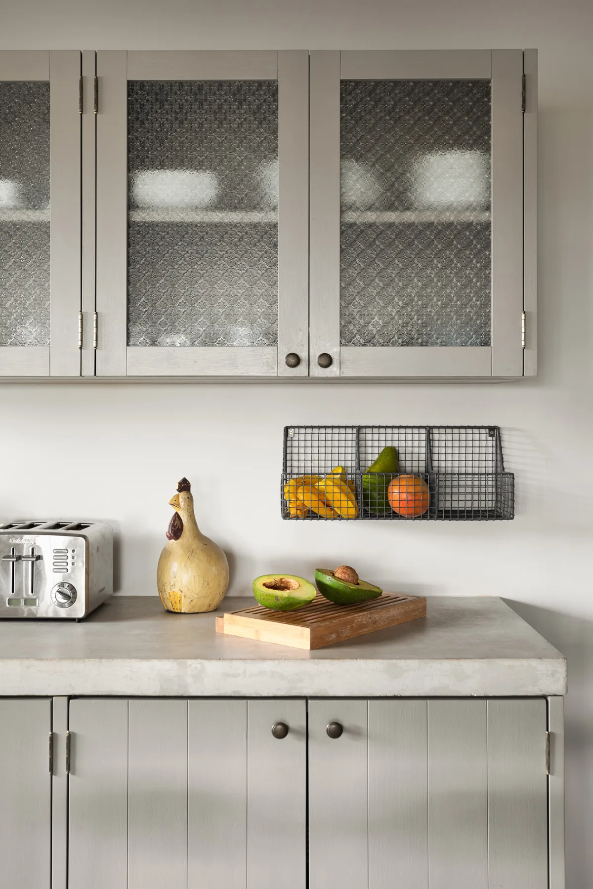 Grey cabinets with textured glass doors hang above a concrete countertop styled with a toaster, wooden cutting board, and a black wire fruit basket.