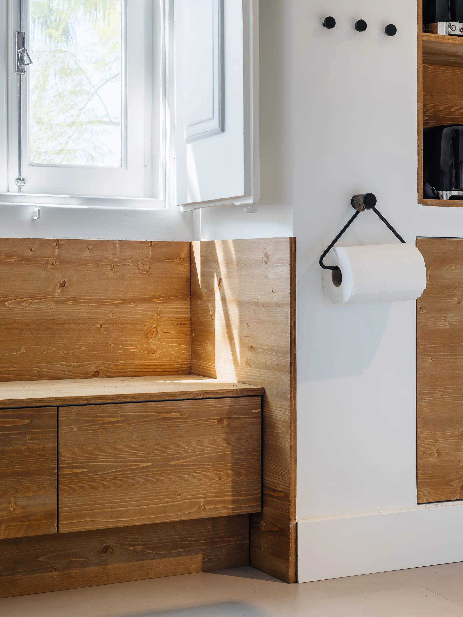 A natural wood built-in storage bench sits below a white window with wood shutters, flanked by minimalist black wall hooks and a paper towel holder.