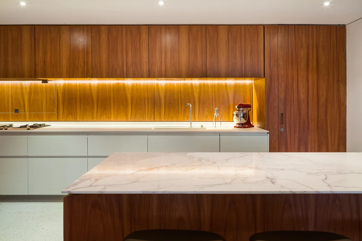 A modern kitchen with a marble-topped island, warm wood upper cabinets, off-white lower cabinetry, and under-cabinet strip lighting.