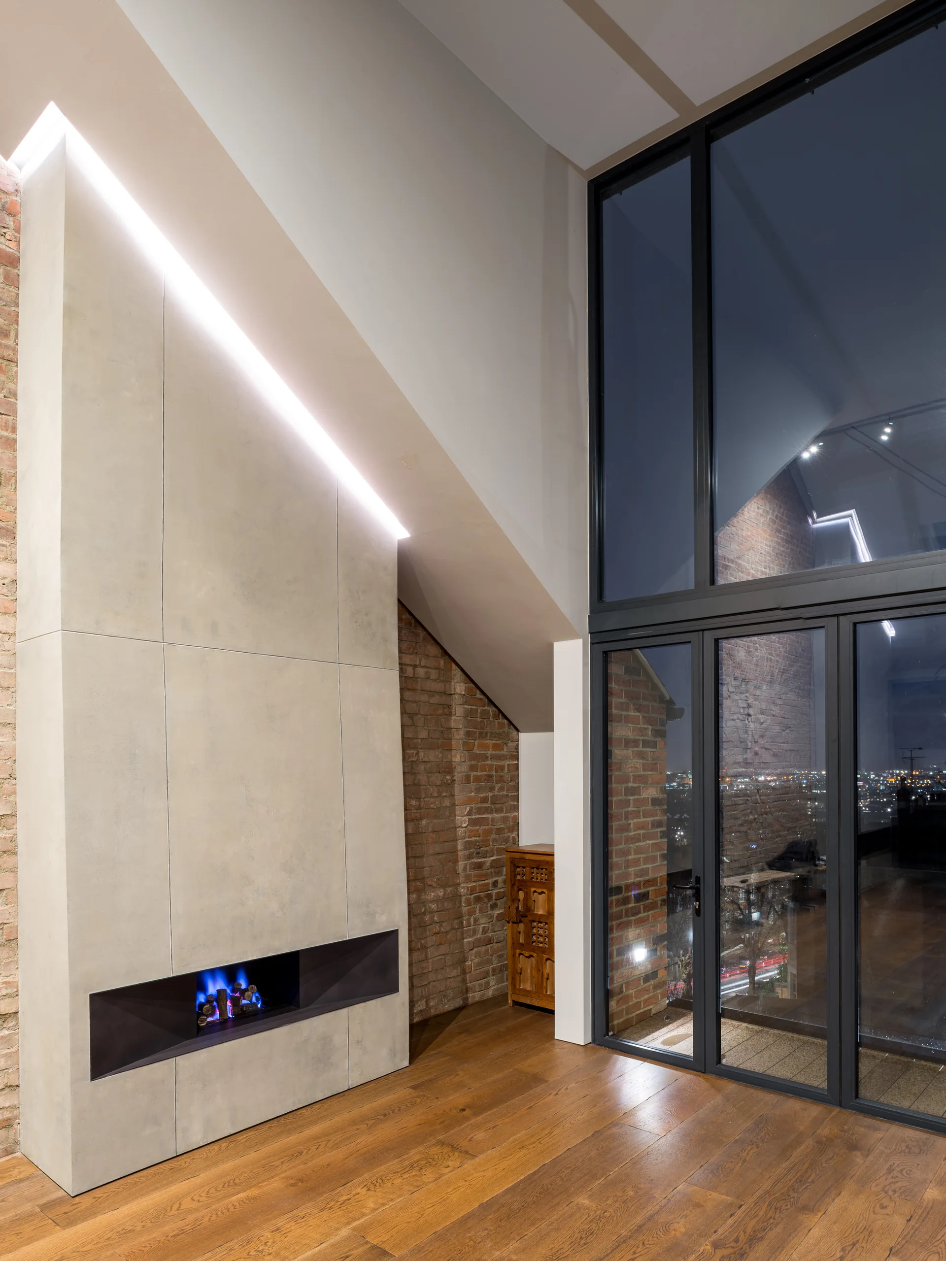 A modern living space featuring a tall concrete-panel fireplace with a linear blue flame beside an exposed brick wall and large glass doors.