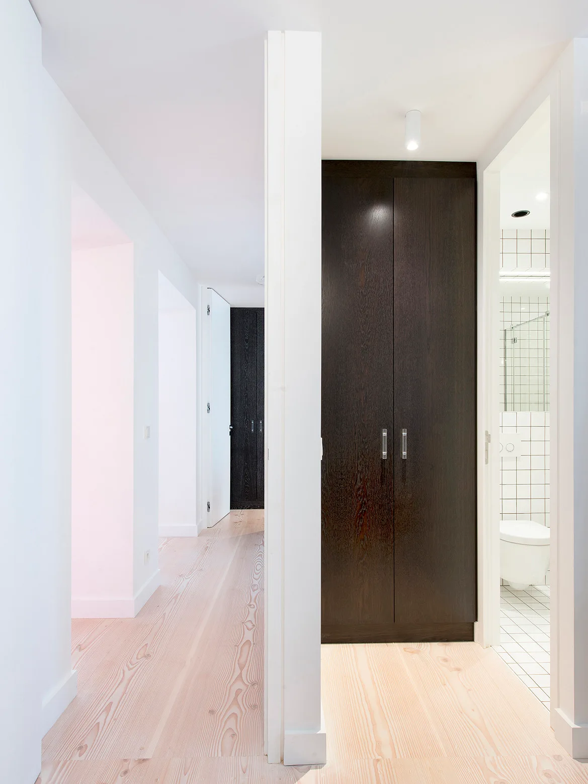 A bright white hallway features light-toned wood floors, dark wood floor-to-ceiling cabinetry, and a view into a bathroom with white square grid tiles.
