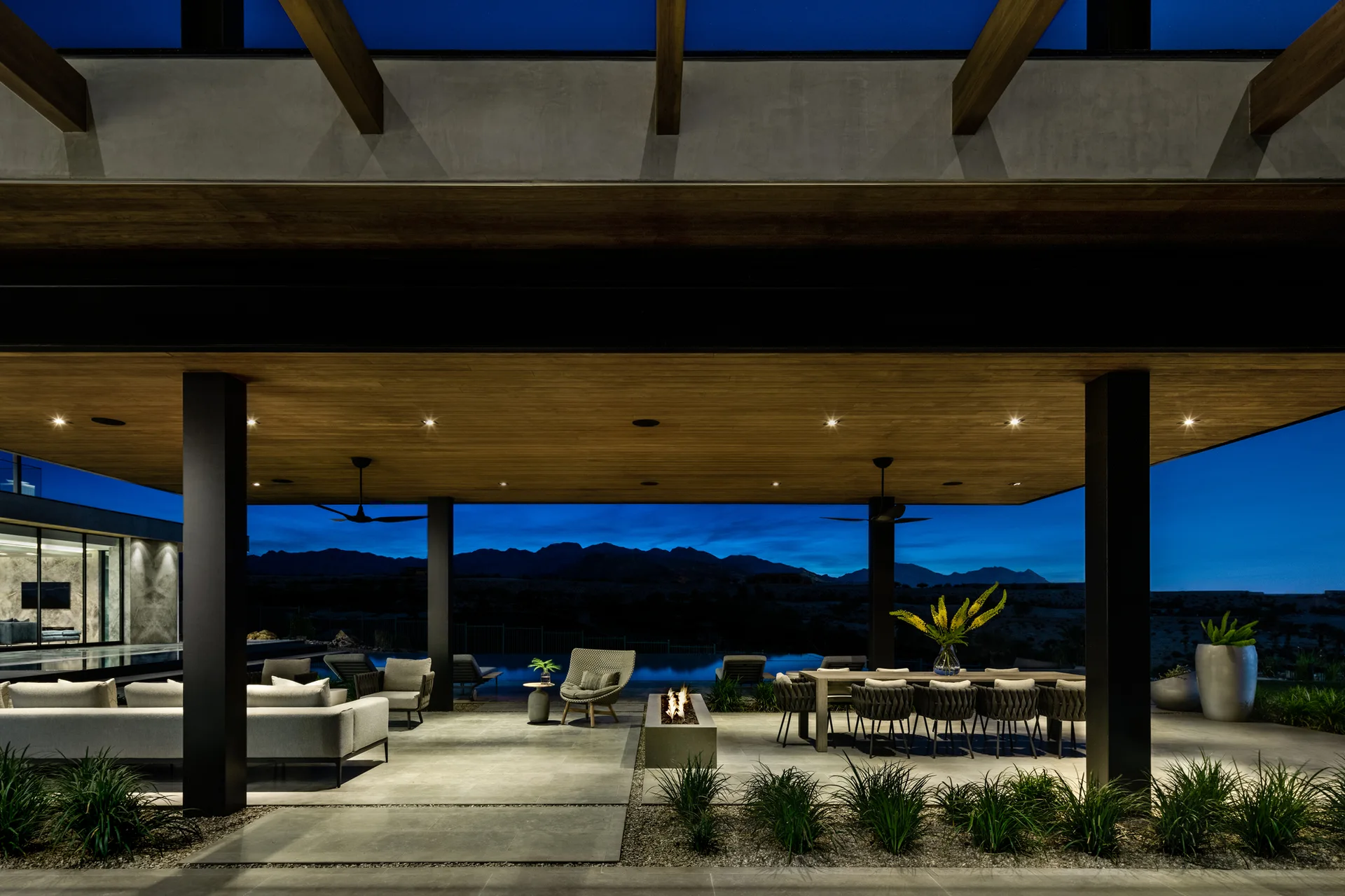 A large covered outdoor patio at twilight features grey sectional seating, a modern fire pit, a long dining table, and a pool with mountain views.